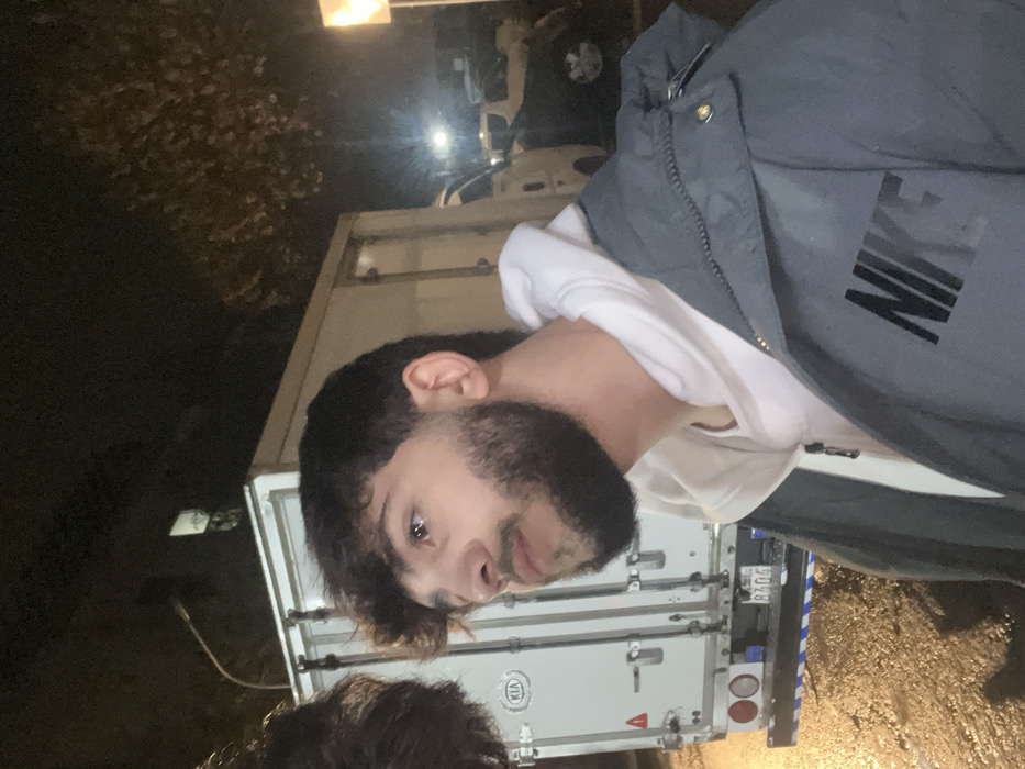 A person with dark hair and a beard is facing the camera, wearing a white hoodie and a gray Nike jacket. Behind them is a white delivery truck with a KIA logo. The license plate is partially visible, showing "8404." The setting appears to be outdoors at night, with streetlights illuminating the scene and foliage visible in the background. The ground seems wet, suggesting recent rain.