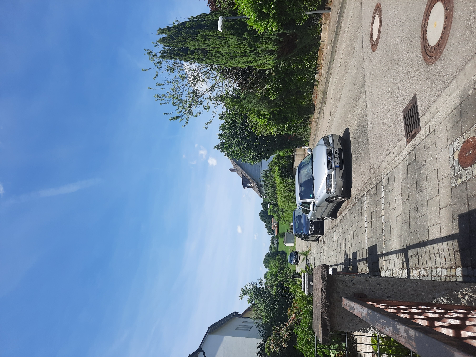 The image captures a street scene in Rosenheim, Germany on a clear, sunny day with a blue sky and wispy clouds. A residential street is lined with houses and lush green vegetation. In the foreground, a gray Volvo S60 sedan with a German license plate "RO-SW 85" is parked on the right side of the road. To the left of the car, a cobblestone sidewalk runs along the street, bordered by a stone wall and railing. Further down the street, a black sedan and another vehicle are visible. The road surface shows two manhole covers and a storm drain. The overall impression is a quiet, suburban environment.