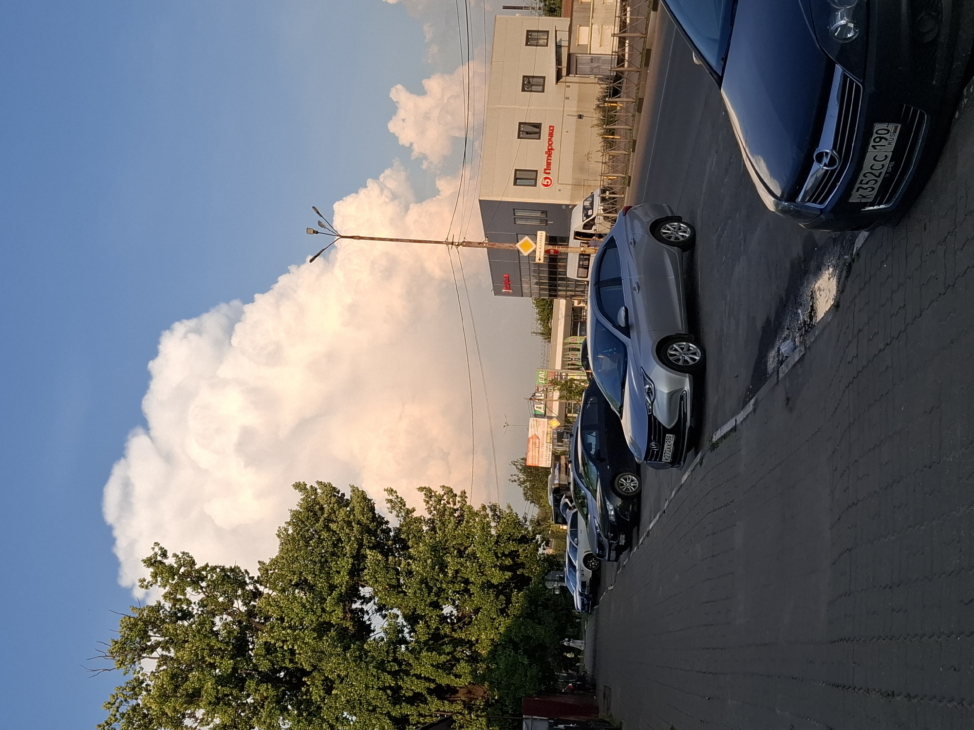 An outdoor vertical, eye-level shot captures a street scene with parked cars and a building under a partly cloudy sky. In the foreground, the dark asphalt and a sidewalk with brick pavers are visible. Several cars are parked along the sidewalk, with a dark blue Opel in the immediate foreground. Further down the street, a silver sedan and other vehicles are parked in a row. To the right, a modern, light-colored building with red lettering that appears to say "Пятёрочка" is situated across the street. A streetlamp with multiple bulbs stands near the building. A large, leafy green tree occupies the left side of the frame, obscuring some of the background. The sky above is a vibrant blue, dotted with large, fluffy white and pinkish clouds, illuminated by the setting sun. The overall scene suggests a typical urban environment during a late afternoon or early evening.