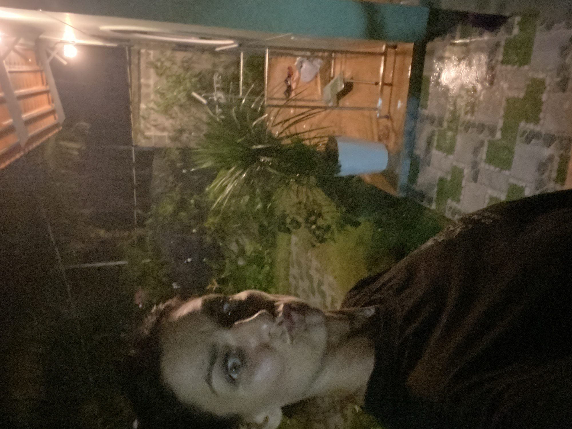 A selfie taken at night on a tiled patio, possibly in Mũi Né, Vietnam. A woman with dark hair and wide eyes is visible in the foreground, puckering her lips. She is wearing a dark brown shirt. Behind her, a porch light illuminates a potted plant and a railing. The patio is wet, suggesting it may have rained. The background is dark and obscured by foliage.
