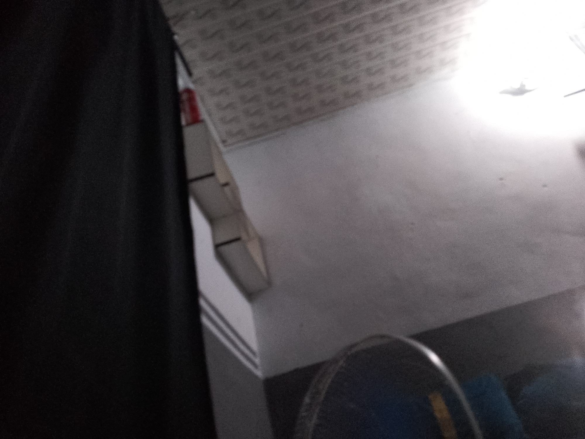 The image depicts an interior scene, potentially a room or hallway, with a ceiling covered in a repeating patterned tile. To the left, a large, dark, out-of-focus curtain or fabric dominates the foreground. In the mid-ground, a white, rectangular object with a red stripe is partially visible, mounted on a wall or shelf structure. Further to the right, a bright light source is visible, casting a strong glare on the wall. At the bottom right of the image, the edge of a dark, circular object, possibly a fan, is visible. The overall lighting is uneven, with a bright glare and darker shadowed areas. The scene is devoid of any discernible people or specific actions. There are no identifiable elements that would definitively place this scene in the city of Jauro Yinu, Nigeria; the description is limited to the visual information present in the image.