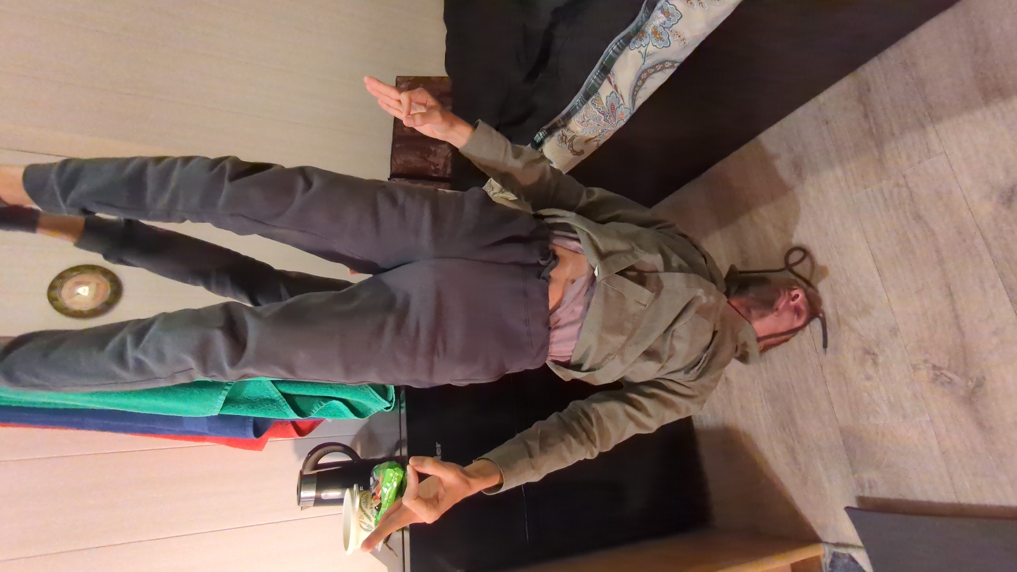 A man wearing grey sweatpants and a tan jacket lies on his back on a wood-look floor. His head is near a wall. His left arm extends toward a wall that has a towel rack with green, blue, and red towels hanging on it. Next to the towels are an electric kettle and a cup. His right arm extends over the end of a dark-colored couch with a decorative cushion. A small bronze-colored decorative plate hangs on the wall. The location is in Gelendzhik, Russia.