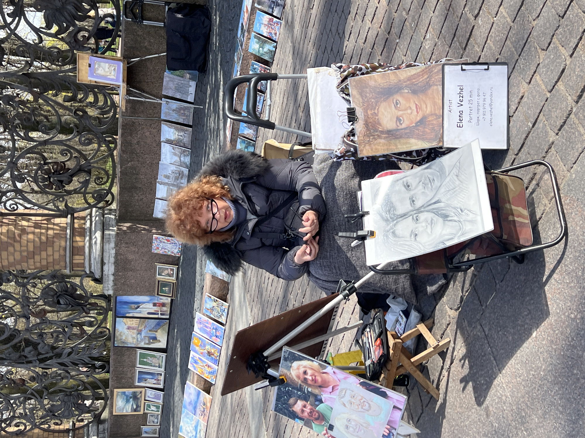 The image shows a street artist, Elena Vezhel, sitting next to her artwork displayed for sale on a paved pedestrian area in Saint Petersburg, Russia. She is wearing a dark winter coat with a fur-lined hood and glasses, and she's holding something small in her hands. Next to her is a small, wooden folding stool.
Her artwork includes sketches, portraits, and paintings. One sign mentions "Portret 25 min" and provides a phone number and email address. The artwork is displayed along a decorative iron fence and on a small rolling cart.