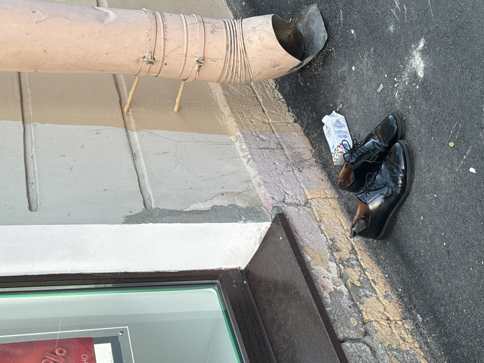 In Saint Petersburg, Russia, a pair of glossy black leather oxford shoes are positioned on a grey asphalt surface near a building's facade. The shoes are situated next to a partially crumpled white carton, possibly for milk or juice, which displays colorful festive imagery. The building features a pink-colored downspout running vertically along its beige and white painted wall. The lower section of the wall exhibits chipped paint and exposed brickwork. To the left of the scene, a dark brown window sill and a portion of a window frame are visible, hinting at the presence of a commercial establishment with a display inside. The overall environment suggests an outdoor urban setting.
