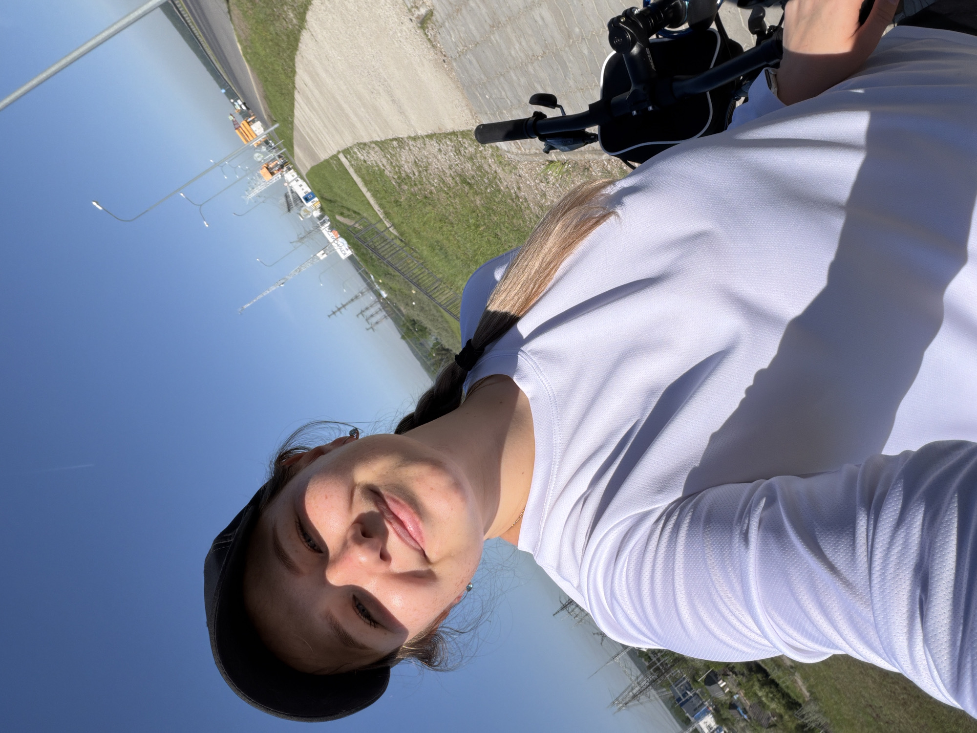 A young woman in a white shirt and dark baseball cap poses for a selfie outdoors. She is seated on a bicycle, with the handlebars visible in the upper right. Behind her, a concrete path and grass verge lead to a road lined with streetlights and parked trucks. In the distance, a body of water is visible. The picture appears to be taken during the daytime under a clear, blue sky. Based on the details present, it is likely this photograph was taken in or around the city of Chaikovsky, Russia.