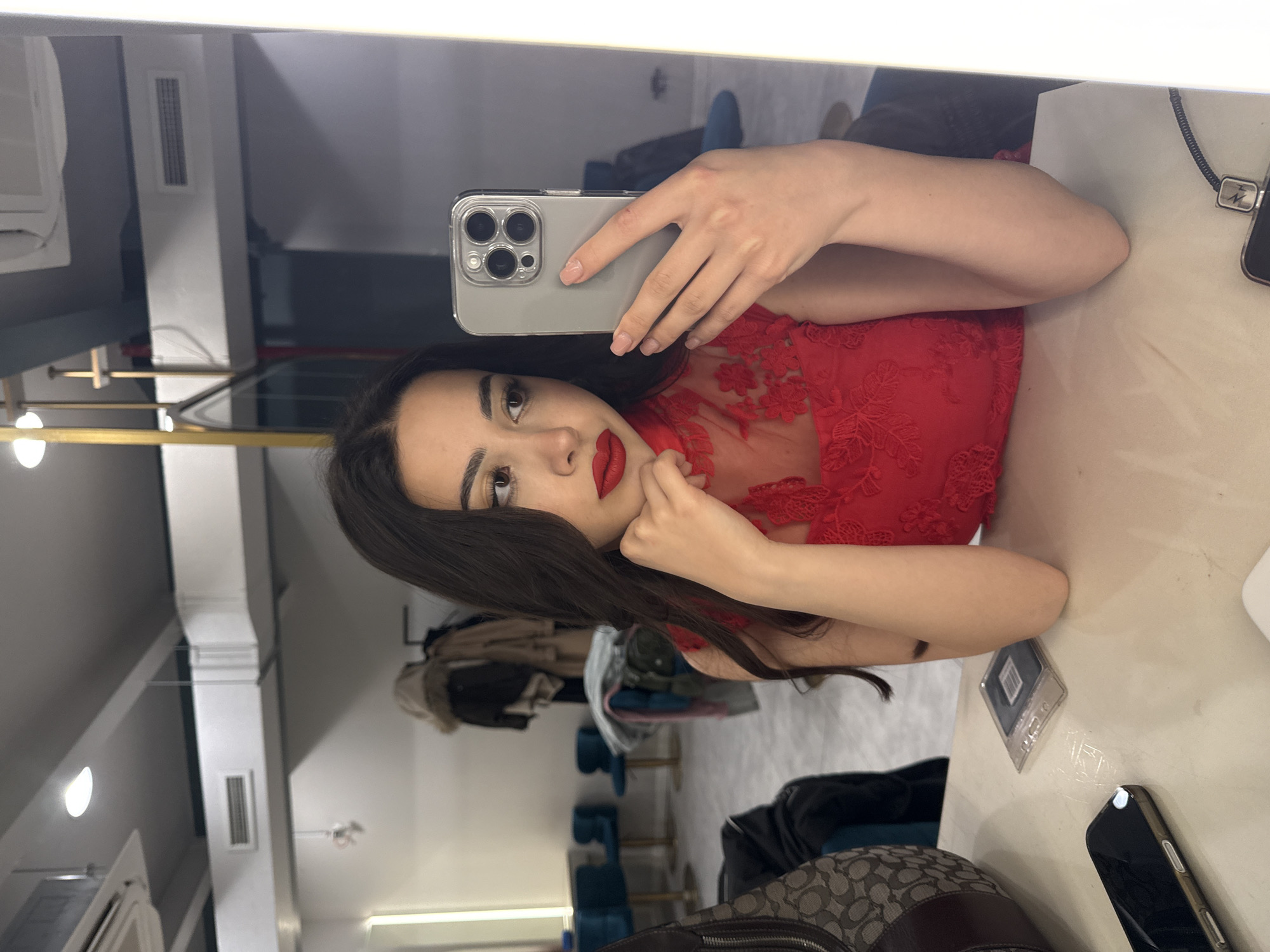 A young woman with dark hair and red lipstick is holding a smartphone (likely an iPhone 13 Pro or 14 Pro) to take a selfie in a mirror. She is wearing a red lace top. The background suggests an interior space with clothing racks and what appears to be a dressing room or retail environment. Based on the presence of Russian language product packaging in Yerevan, Armenia. The scene also includes a counter or table with a mobile phone and a purse on it. The lighting appears to be artificial, indicating an indoor location.