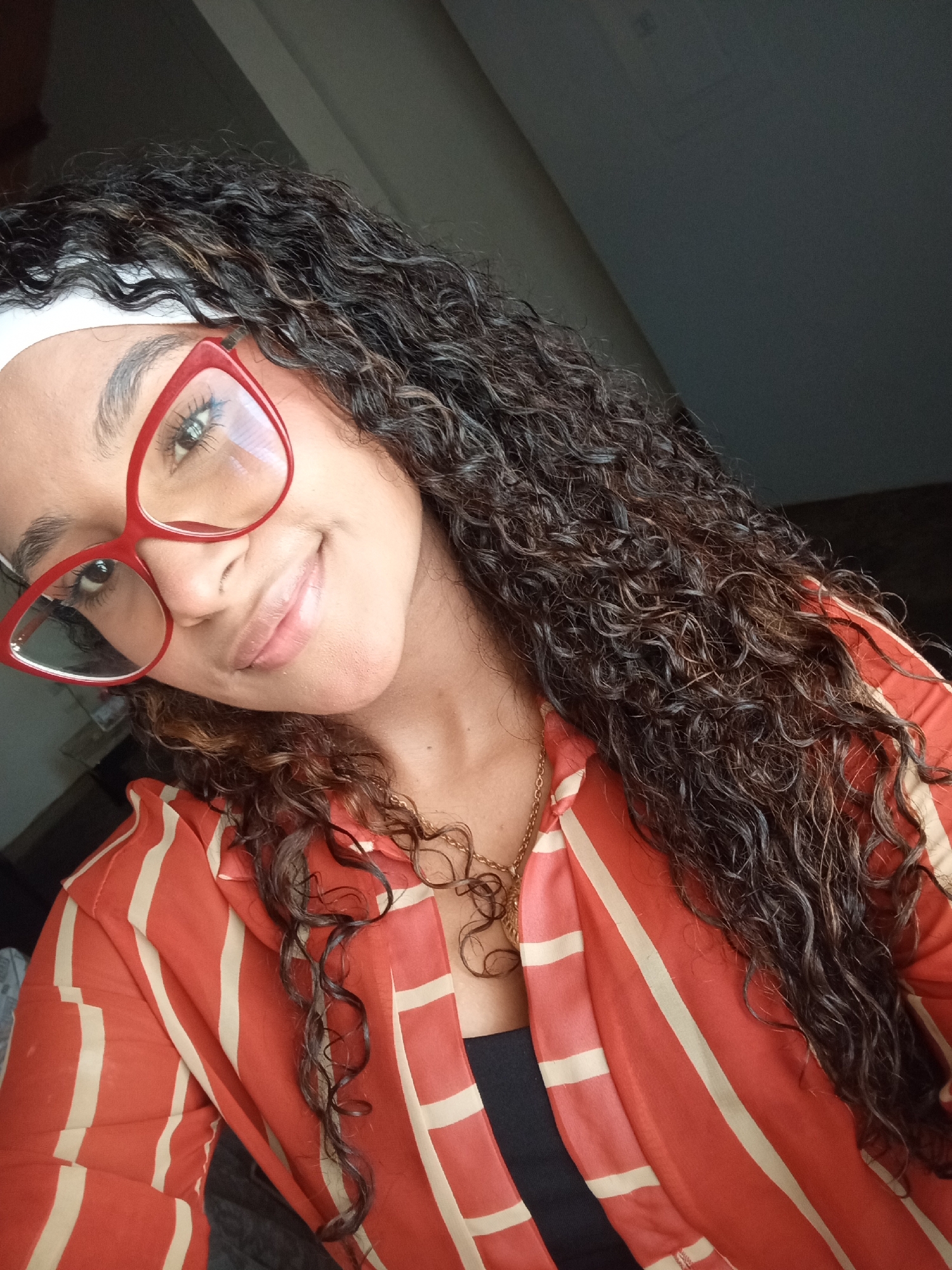 A woman with curly brown hair and a white headband is wearing red glasses and a red and white striped shirt. She is smiling slightly and wearing a gold necklace. The background is dark, possibly indoors. There is no information available about whether or not the image was taken in Juan Griego, Venezuela.