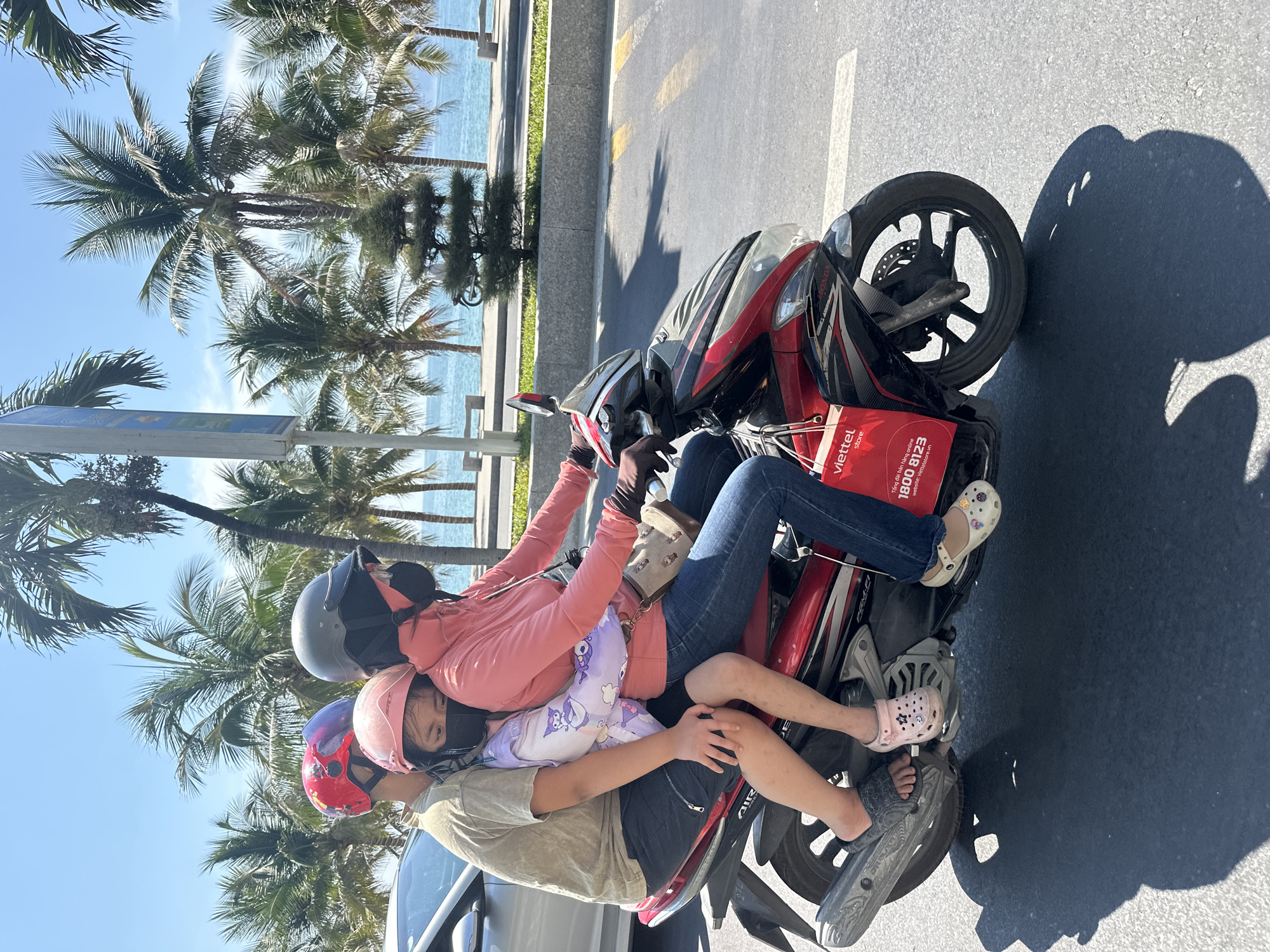 The photograph shows three individuals on a red and black motorcycle, with a coastal scene featuring palm trees and the ocean in the background. The location appears to be Nha Trang Ward, Vietnam.
The person in front is wearing a gray helmet, pink jacket, and blue jeans, operating the motorcycle. A medium tan purse hangs off of the left shoulder. The middle individual wears a pink and red helmet and sits in front of the operator, and the third individual sits on the back of the motorcycle, wearing a red helmet and a tan shirt.
The motorcycle is riding on a paved road, and there is a red Viettel bag attached to the back of the seat. The background includes palm trees lining the coast and the ocean under a clear sky. Shadows suggest bright daylight.