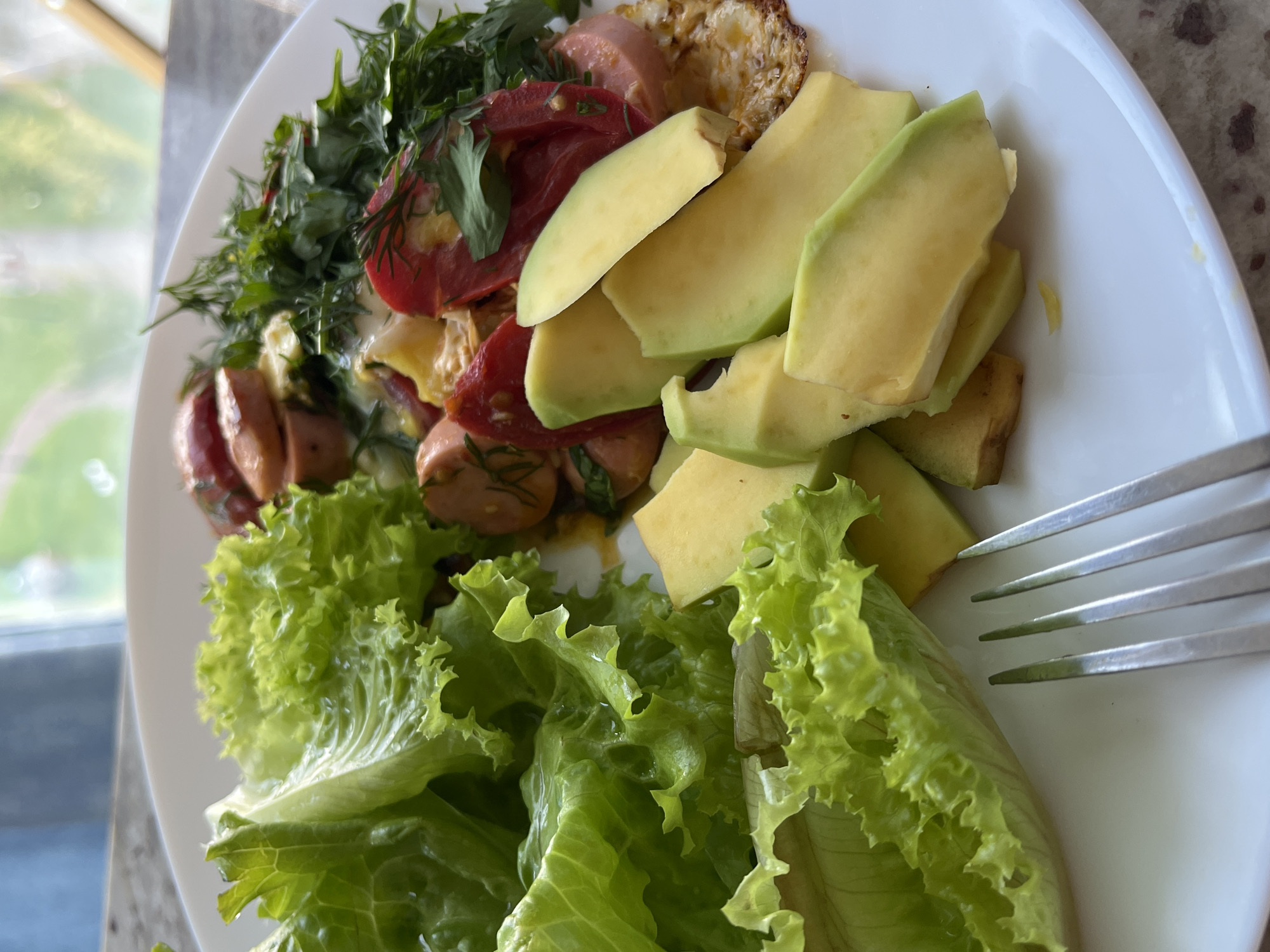 A close-up, high-angle shot captures a white plate of food on a light-colored, speckled surface, likely a table. The food consists of a generous portion of bright green, ruffled lettuce on the left, followed by sliced avocado in the center. To the right of the avocado, a cooked egg is visible, topped with sliced sausages, red tomato wedges, and a sprinkle of fresh green herbs, possibly dill and parsley. A silver fork with four tines rests on the lower left edge of the plate. The background is blurred but suggests an indoor setting with a window offering a view of greenery and possibly a building. The lighting is natural, coming from the window, and casts soft shadows. The scene appears to be in an urban setting, possibly a residential apartment or a cafe, given the window view.