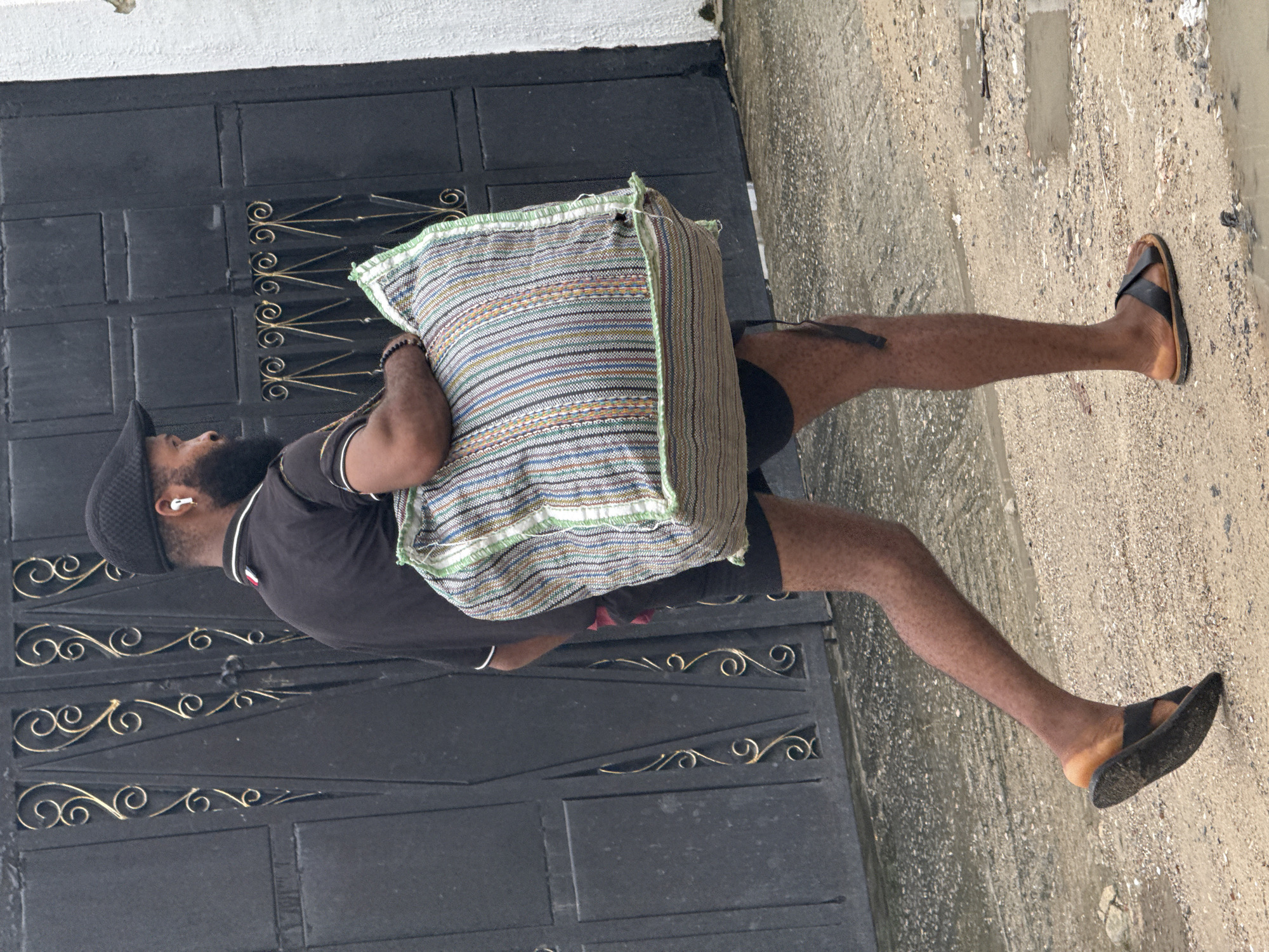 In Ado, Nigeria, a man with a beard, wearing a black cap, black polo shirt, and black shorts, walks on a dirt and gravel path. He carries a large, multicolored woven bag over his left shoulder. The man is also wearing black flip-flops. He has white earbuds in his ears. Behind him is a black metal gate with decorative gold scrollwork. To the right of the gate is a white wall. The ground appears wet in places, suggesting recent rain.