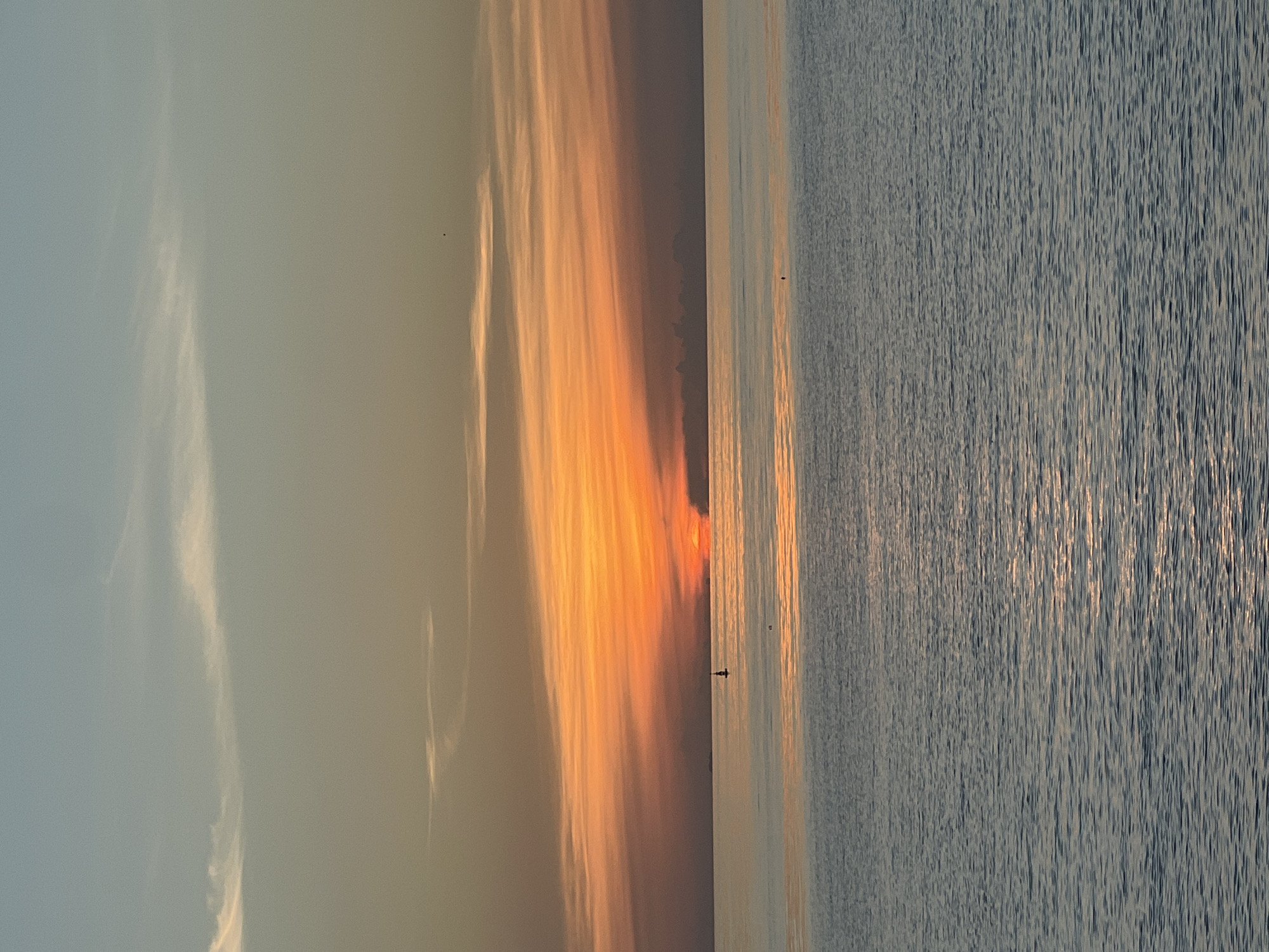 A serene sunset over the water is captured in this vertical, eye-level shot. The horizon line divides the image, with the bottom two-thirds filled with calm, rippling water that reflects the warm hues of the sky. The water's surface is a deep blue-grey, with shimmering highlights indicating the sun's rays.
The top third of the image is dominated by the sky, which transitions from a pale blue at the very top to a vibrant orange near the horizon. Streaks of thin, wispy clouds are scattered across the sky, catching the sunset light and creating a textured effect. The sun itself is partially obscured by a band of darker clouds along the horizon, appearing as a bright orange orb with its lower half hidden.
In the middle distance of the water, a solitary black buoy with a white top is visible, marking a point in the vast expanse. Another small, dark dot, possibly a distant boat or a bird, can be seen in the upper right quadrant of the sky. The overall mood is tranquil and peaceful, typical of a sunset over a large body of water. The location is the coast of Ko Pha-ngan District, Thailand, likely viewed from a beach or a boat.