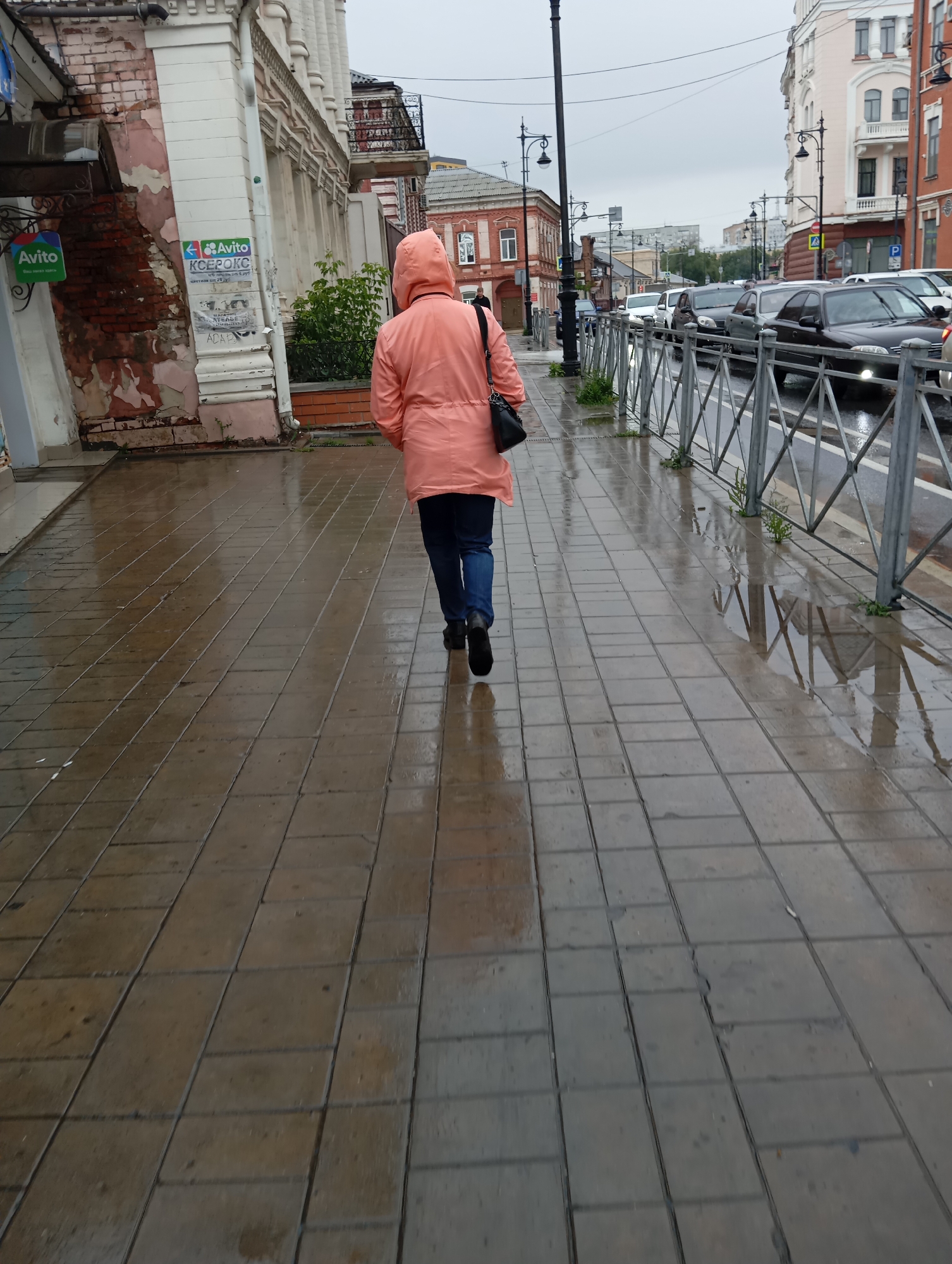 A person in a light pink hooded jacket and blue jeans walks away from the camera down a wet, tiled sidewalk in Orenburg, Russia. The person is carrying a black purse over their right shoulder. The sidewalk is reflective due to the rain. To the left of the sidewalk, a dilapidated building with exposed brick and peeling paint is visible. A sign with the "Avito" logo is attached to the building. To the right of the sidewalk, a metal fence separates the sidewalk from a street where several cars are driving in traffic. The sky is overcast, indicating rainy or cloudy weather. The architecture visible in the background suggests an urban setting with older buildings.
