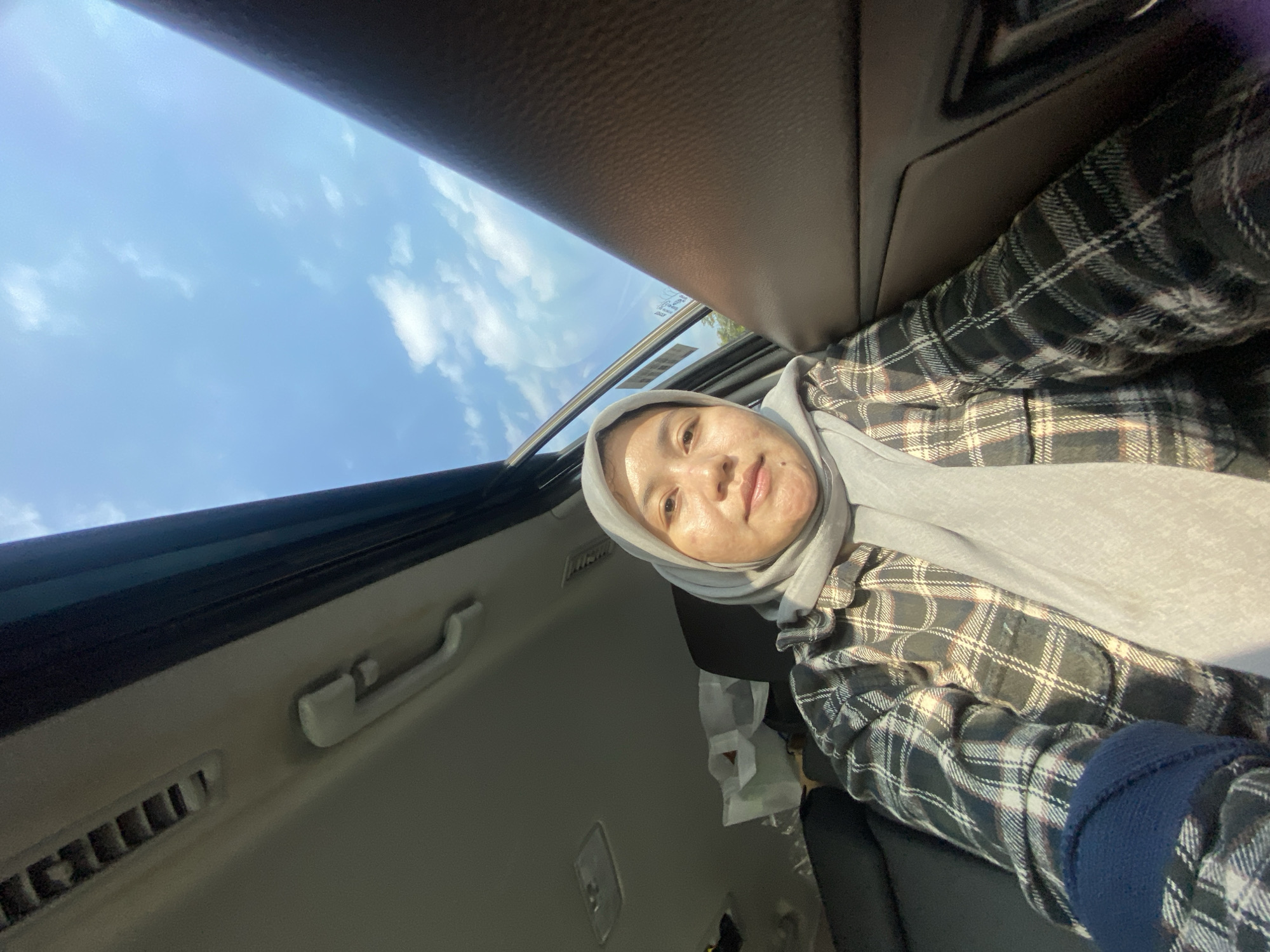 A young woman wearing a grey hijab and a plaid shirt is inside a vehicle, looking directly at the camera. The interior of the car is visible, with the ceiling and part of the dashboard and door panel. Outside the vehicle, a clear blue sky with scattered white clouds is visible through what appears to be a sunroof or an open window. The woman's facial features are clear, and she has light skin with some visible blemishes. The lighting suggests it is daytime. The image does not provide any specific visual cues that would allow for identification of the city of Banjarmasin, Indonesia.