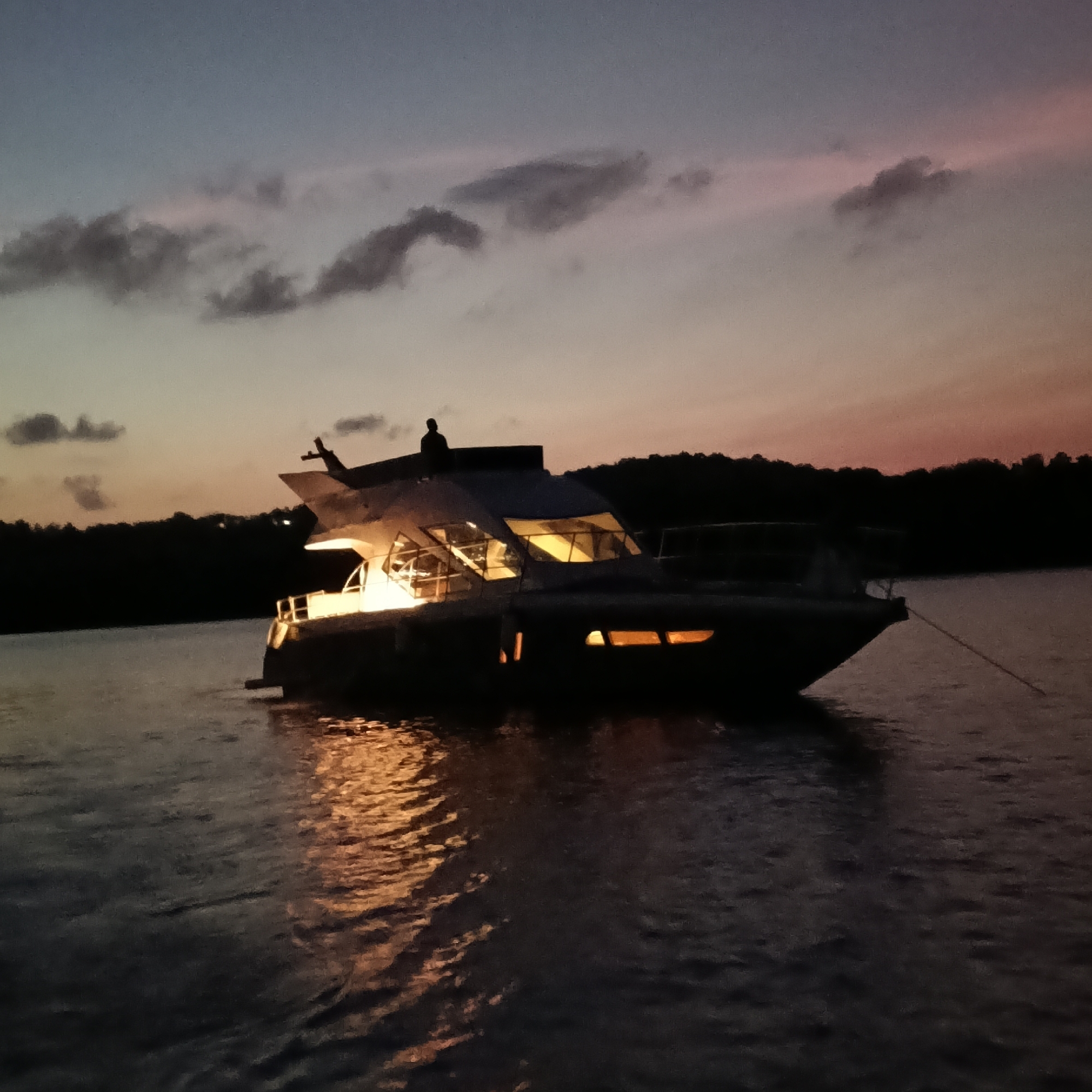 A yacht sits anchored on a body of water, with dark trees silhouetted on the far shore under a dusky, pink-tinged sky with scattered clouds. Lights emanate from the yacht, illuminating the surrounding water. A person appears to be standing on the top deck of the yacht. A rope extends from the bow of the yacht into the water. The location is likely not Britona, India as it is a small village and this scene is not typical there. The image contains a yacht, water, trees, sky, clouds, person, lights, and rope. The time is likely dusk or dawn.