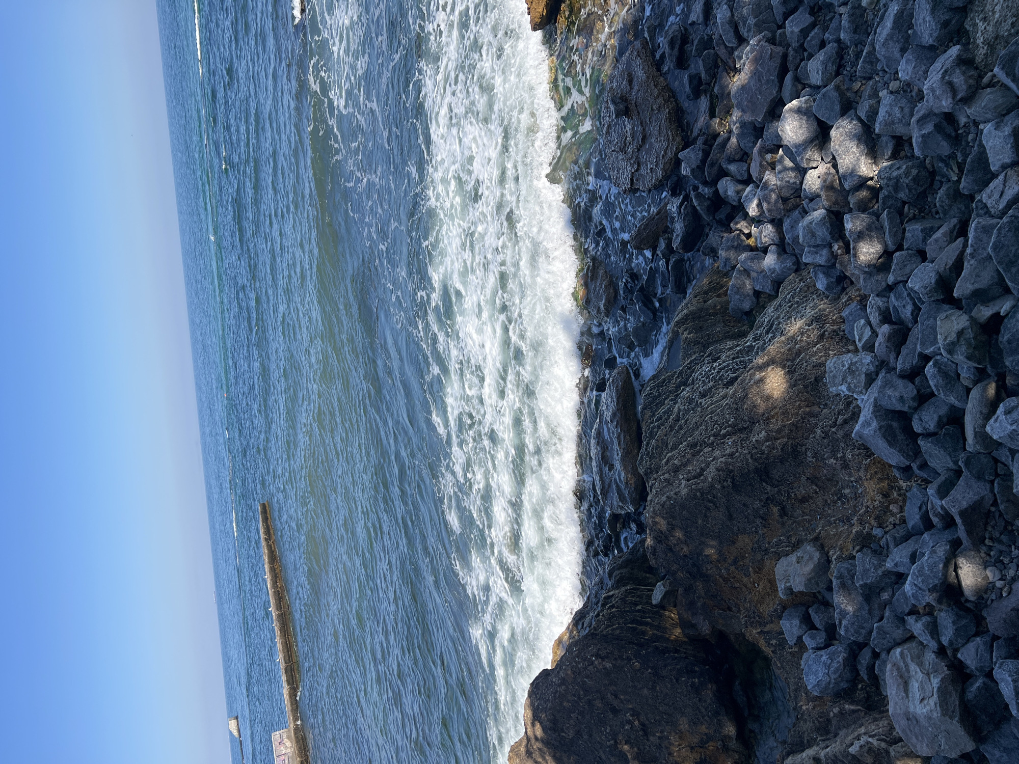 The image shows a rocky shoreline and the sea under a clear blue sky. In the foreground, there are many large and small dark gray rocks. To the left, a large, dark, textured boulder casts a shadow on the rocks in front of it. In the midground, waves crash against the shore, creating white foam. Further out, the deep blue sea stretches to the horizon, with a faint line of a distant object, possibly a ship or structure, visible in the upper left quadrant. There is a concrete pier extending into the water from the left side of the image, with several white birds perched on it. The overall scene depicts a natural, coastal environment.