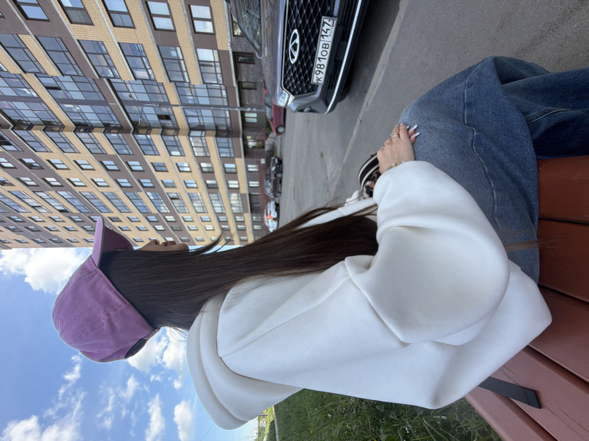 The image captures a young person sitting on a brown wooden bench in an urban outdoor setting during the daytime. The person, viewed from a side-back perspective, has long dark hair and is wearing a white hooded sweatshirt, blue denim jeans, and a purple baseball cap. Their hands are visible, with one resting on a knee, showing long, manicured nails with a light ombré design, along with several rings and a bracelet.
In the background, a large, modern apartment building with a light beige and brown facade and numerous windows stretches upwards. A bright blue sky with scattered white clouds is visible. Parked on a paved surface next to the bench is a dark grey SUV. Its front grille, featuring a Chery logo, and license plate are clearly discernible. The license plate reads "K981OB 147 RUS," indicating Russia and a Moscow region code. Other vehicles, including a red sedan, are visible further down the street. Green grass and some foliage are present in the foreground and around the building. The scene depicts a relaxed moment in what appears to be a residential area in a Russian city.