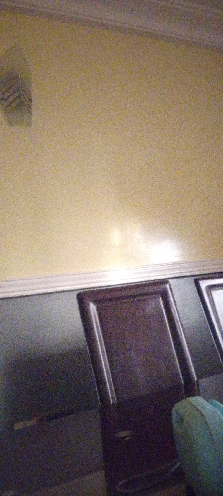 The image shows an interior scene, possibly a room. The wall is painted a pale yellow color and has a white crown molding at the top. Below the molding is a dark gray surface, which could be a wall, a cabinet, or a piece of furniture.
In the foreground, there is a dark brown chair with a carved panel. To the right of the chair, a portion of another chair or furniture piece is visible, with a lighter color. On the far right, a light blue object, possibly a cushion or a bag, is partially visible.
On the far left of the image, near the top of the pale yellow wall, there is a light-colored object mounted on the wall. It appears to be a square or rectangular fixture with angled slats or grooves.
The image is somewhat grainy and appears to be taken in low light conditions. There are no people visible in the frame. The overall impression is that of a sparsely furnished room. The location in Madalla, Nigeria, is not explicitly depicted by any landmarks or context within the image itself.