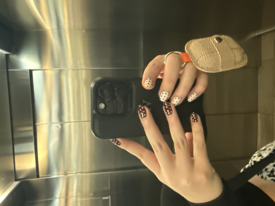 An individual is taking a mirror selfie inside an elevator in Mexico City, Mexico. The subject holds a black-cased smartphone, identified as an iPhone with a triple-lens camera system, centered in the frame. Faint "LV" branding is visible on the back of the phone case. The left hand's fingernails are painted dark brown with white polka dots. The right hand's fingernails are painted white with dark brown polka dots and are also holding a keychain attached to a metal ring. The keychain features an orange strap and a beige, textured fabric pouch with a dark button closure. The subject's left arm is partially visible, clad in a dark, long-sleeved garment, and a segment of patterned clothing is visible on the lower left. The elevator interior consists of brushed stainless steel panels, reflecting ambient warm light and displaying minor surface imperfections.