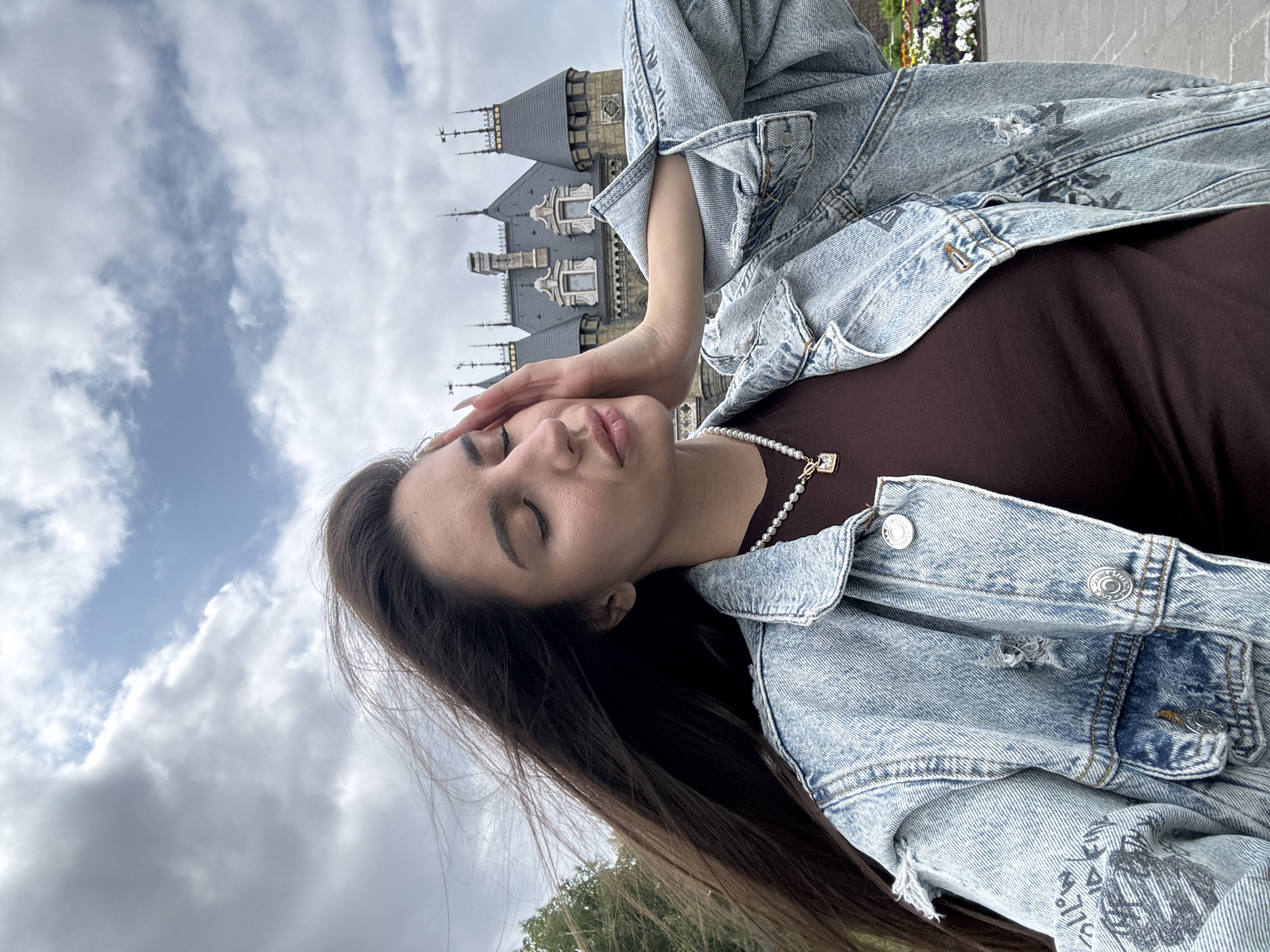 A young woman with long dark hair, wearing a denim jacket and a brown top, is pictured in front of a castle-like building under a cloudy sky. Her eyes are closed, and her right hand is gently touching her temple. The castle, located in Khryashchevka, Russia, has a distinctive architectural style with spires and multiple windows. The foreground shows a stone pathway and some colorful flowers in the background. The woman's hair is blowing in the wind.