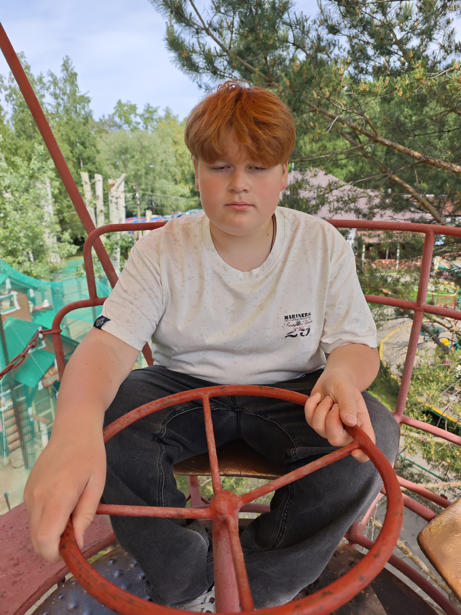 A full shot captures a young male with red hair seated within an amusement park ride. The ride appears to be elevated, offering a view of greenery and structures in the background. The individual is fair-skinned and wears a white short-sleeved t-shirt with a logo on the chest, along with dark-colored jeans. He is holding onto a red metal steering wheel. The ride's framework is also red. The location is identified as the city of Chaikovsky, Russia.