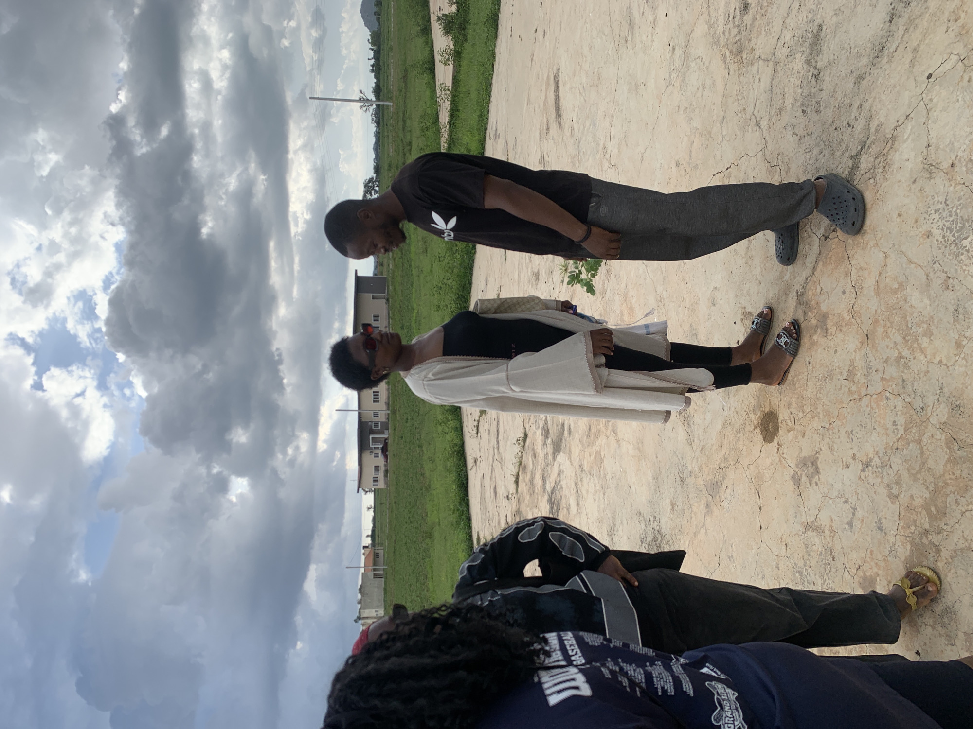 The media file depicts a group of individuals outdoors in Gwafan, Nigeria. The setting appears to be an open, paved area with grassy fields and some buildings in the background. The sky is filled with dramatic, cloudy formations, suggesting overcast or potentially stormy weather.
The subjects include at least three people. In the foreground, a person wearing a dark blue shirt with white text and intricate graphics is partially visible. Behind them, another person is seen from the waist down, wearing dark pants and black and grey patterned outerwear, with one foot in a yellow sandal. Further back, two people are more fully visible, facing each other. One, wearing a black t-shirt and grey pants with grey crocs, looks down. The other, wearing a black top and a cream-colored garment over it, with red-framed sunglasses, looks up.
The activity seems to be a casual gathering or interaction among the individuals. The image is taken in daylight, given the illumination. The cloudy sky creates a somewhat dramatic atmosphere. The architecture of the buildings in the background hints at a residential or developing area. There are no overt signs of a specific event, but the poses suggest conversation or a shared moment.
Visible text on the shirt in the foreground includes "GRAND" and other words that are difficult to discern clearly due to the angle and resolution. The overall impression is one of everyday life in an outdoor Nigerian environment under a dynamic sky.