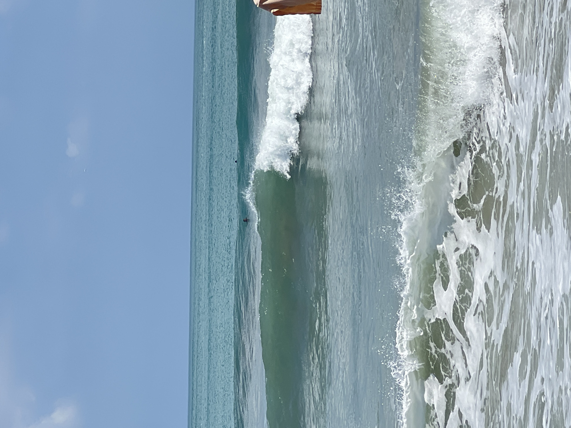 The image shows a beach scene in Kuta, Indonesia. The sky is a clear blue with a few wispy clouds. The ocean is a beautiful turquoise color, with several waves rolling in. One wave is breaking in the foreground, creating a white spray of foam. In the distance, a few people can be seen swimming or surfing in the water. The overall impression is one of a peaceful and idyllic beach day.