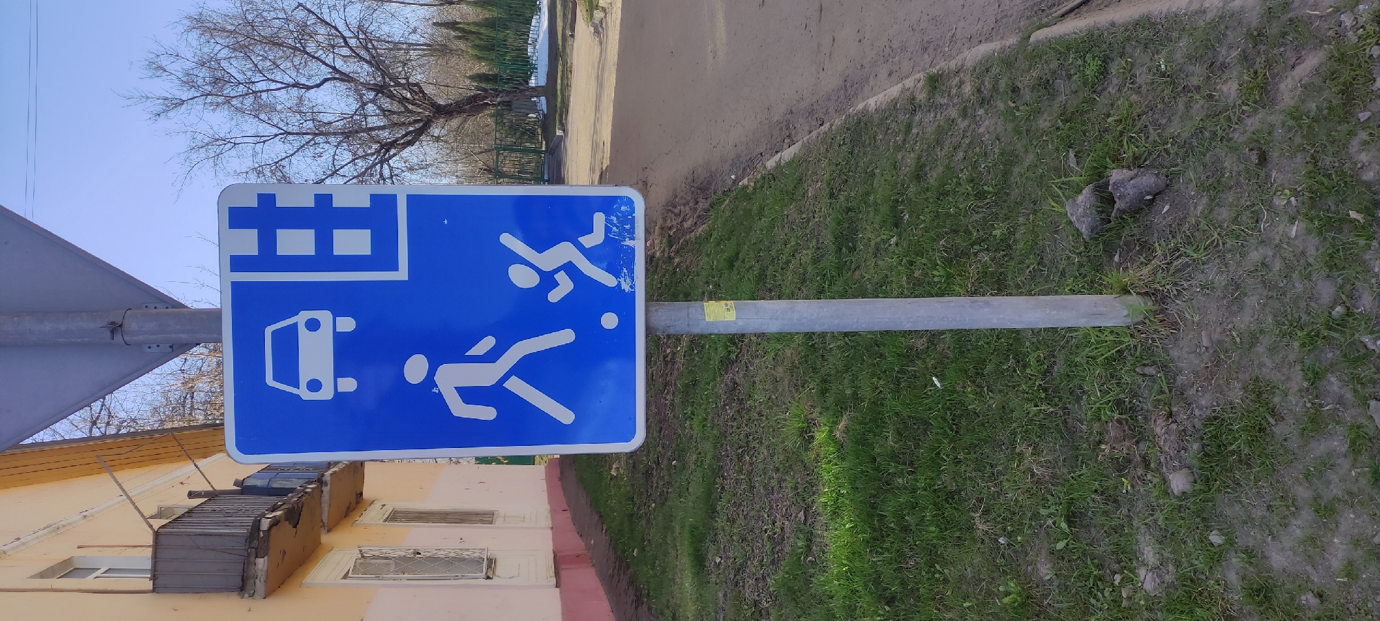 The image depicts a blue and white street sign in Fryazino, Russia. The sign indicates a residential area with children playing. It is mounted on a gray metal pole, rooted in a grassy area beside a sidewalk. Adjacent to the sidewalk is a building with light-colored walls and windows. In the background, a tree with bare branches is visible against a blue sky. A paved road is seen adjacent to the grassy area and sidewalk.