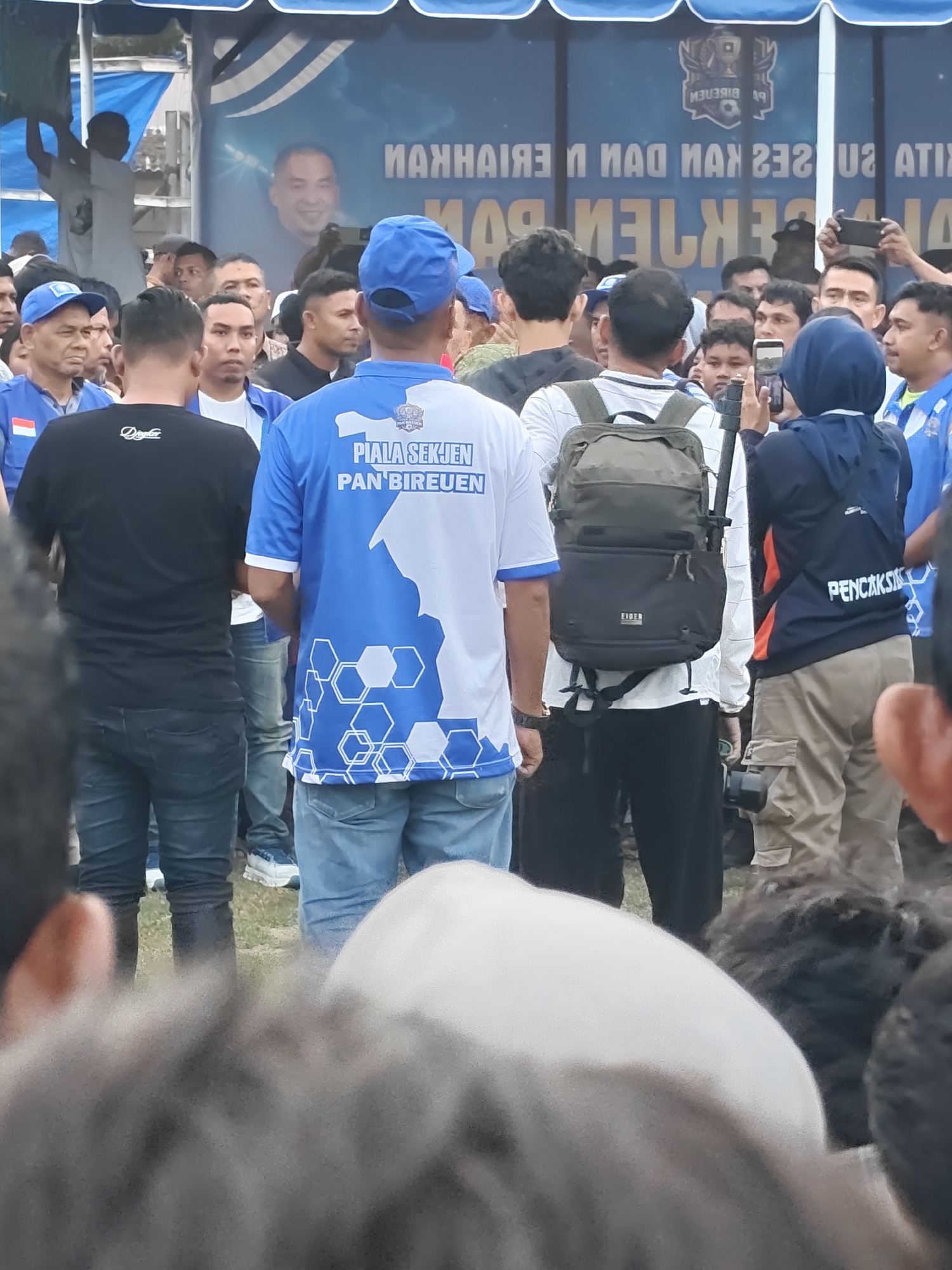 In the city of Desa Meunasah Blang, Indonesia, a crowd of people are gathered outdoors, with a large blue and white tent providing shade overhead. The tent has a banner with text that appears to be in Indonesian and a photo of a man. In the foreground, the backs of several people are visible, most of whom are wearing blue and white shirts, suggesting a common affiliation or event. One person in the center is wearing a blue shirt with white hexagonal patterns and text that reads "PIALA SEKJEN PAN BIREUEN". Another person to their left wears a black t-shirt. A person to the right is wearing a dark blue shirt with orange accents and the word "PENCAKSI" visible. A backpack is prominent on the back of one individual. Several people are holding phones, possibly taking pictures or videos. The ground appears to be covered in grass. The overall atmosphere suggests a community gathering or event.