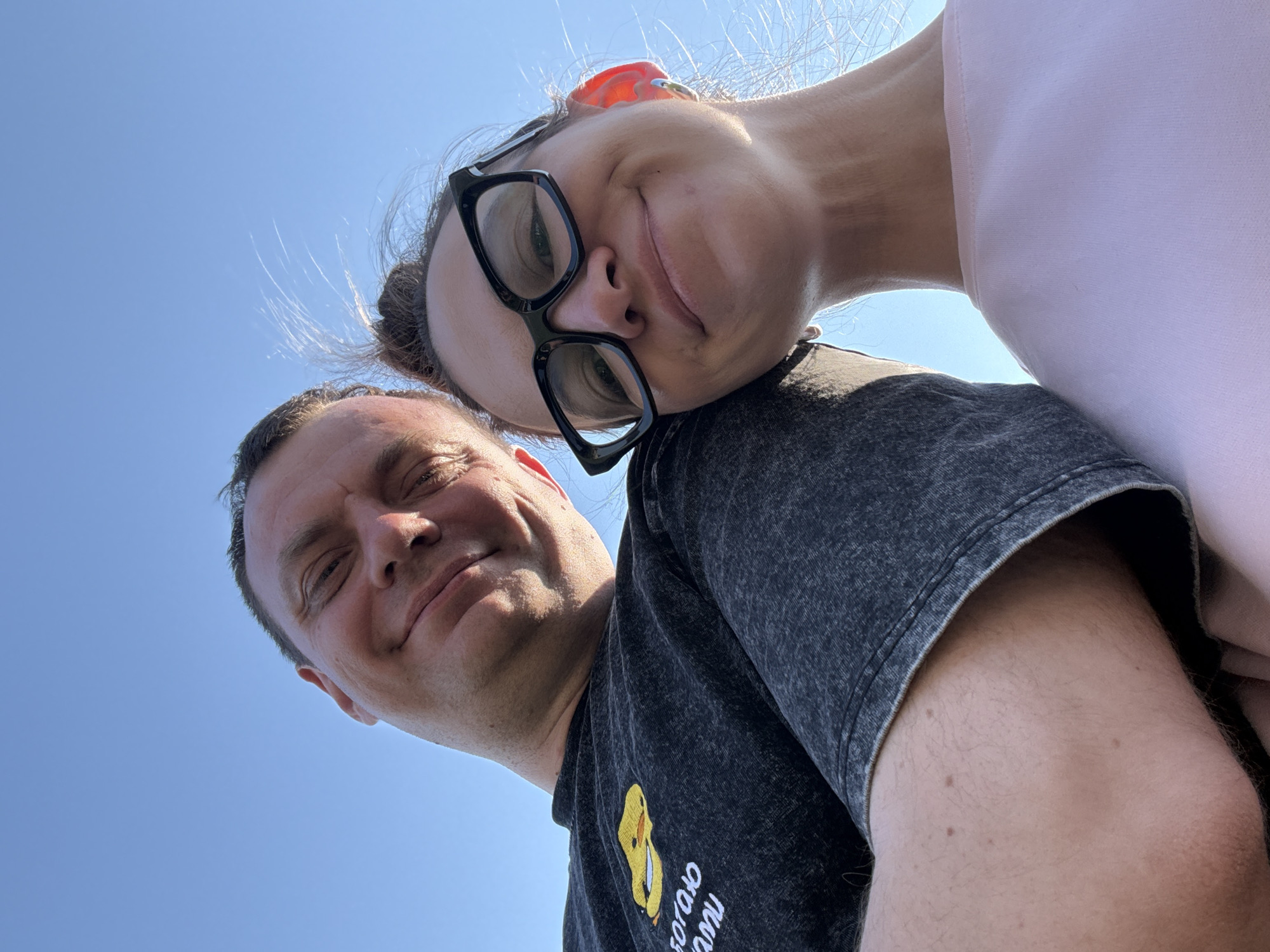 Two people are in the frame, shot from a low angle against a bright blue sky. The woman, wearing glasses and a pink top, is positioned above and slightly behind the man. She has her head tilted to rest against his shoulder. The man is wearing a dark gray t-shirt with a yellow cartoon duck on the lower front left. The picture was taken outside in clear sunny weather. There is no evidence to suggest the location is Kharnavy, Russia.