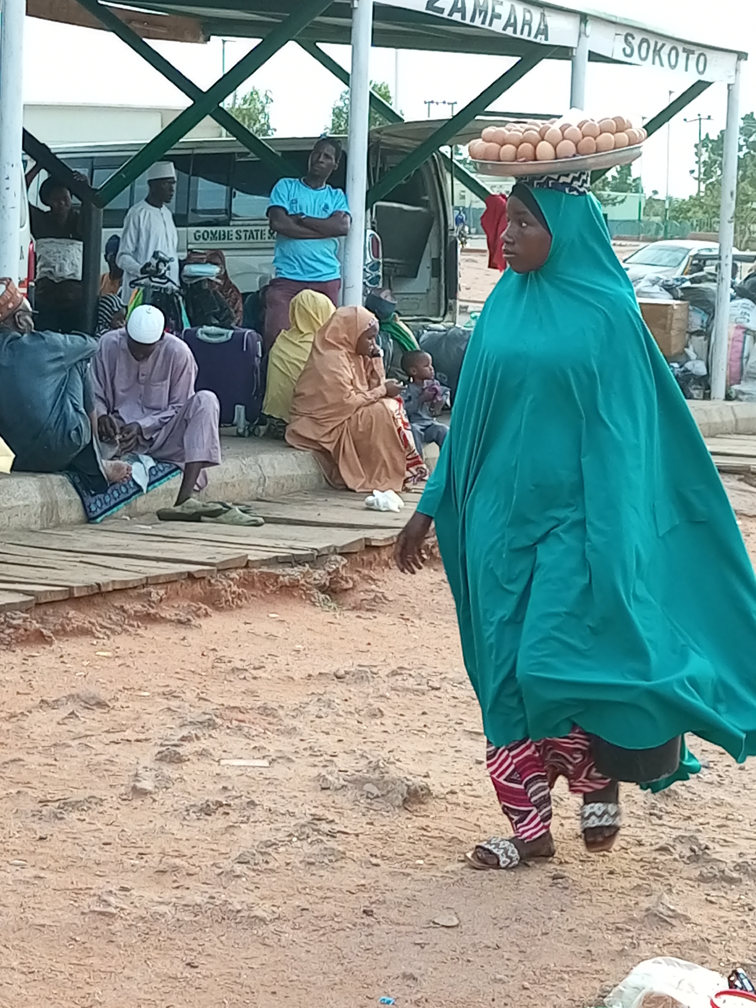The image depicts a street scene in Gombe, Nigeria. In the foreground, a woman wearing a green hijab and patterned trousers walks towards the right, carrying a tray of eggs balanced on her head. Behind her, several people are seated on a raised platform and on the ground. A man in a white cap and gown is seated on the ground, and another man in a blue t-shirt stands with his arms crossed. Luggage is visible around the seated individuals. In the background, a bus is parked, and signs indicate "ZAMFARA" and "SOKOTO". The ground is covered in dirt and sand.
