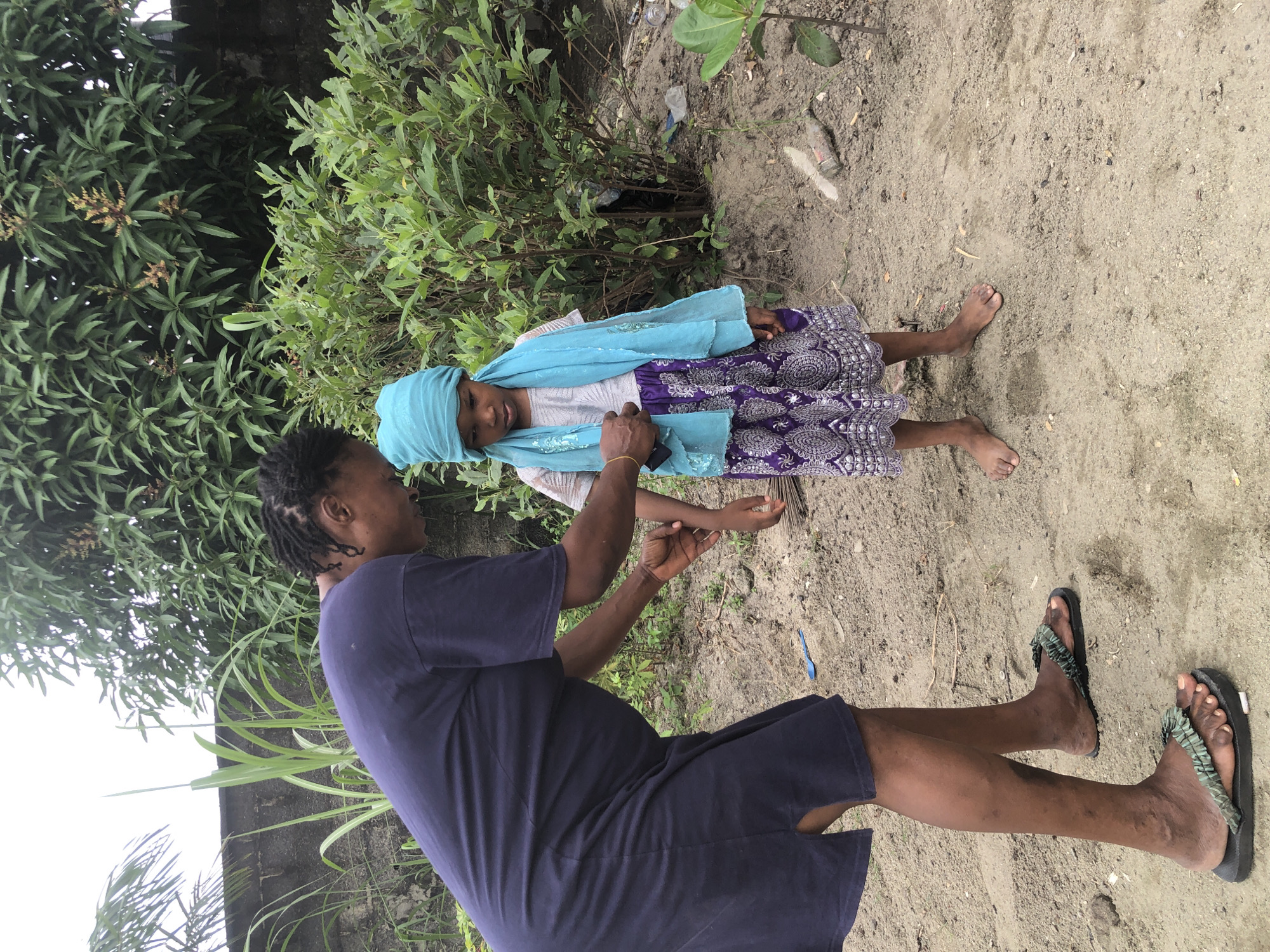 Two people, an adult and a child, are in a sandy outdoor setting with green foliage in the background. The adult, positioned in the foreground and facing away from the camera, is wearing a dark blue t-shirt and dark shorts, with dark sandals on their feet. They have dreadlocks and are holding the child's arm. The child, standing and facing the adult, is wearing a light blue headscarf, a light-colored top, and a purple skirt with a white pattern. The child appears to be looking down and touching their hand. The scene suggests a warm, possibly overcast day, given the general lighting. The presence of vegetation and sandy ground hints at a rural or less developed area. The interaction between the adult and child seems gentle and attentive. There is no visible text in the image.
