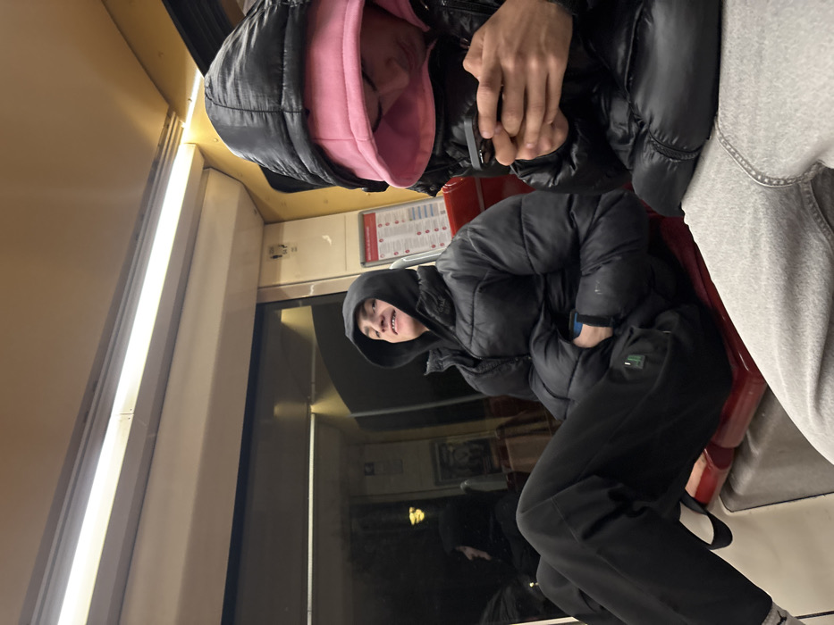 Two people are on a train. One person wears a black puffer jacket with a pink hoodie underneath. The other wears a black puffer jacket with black pants and white sneakers. They sit next to a train window. The train's interior includes red seats and lights. There is a safety information sticker.