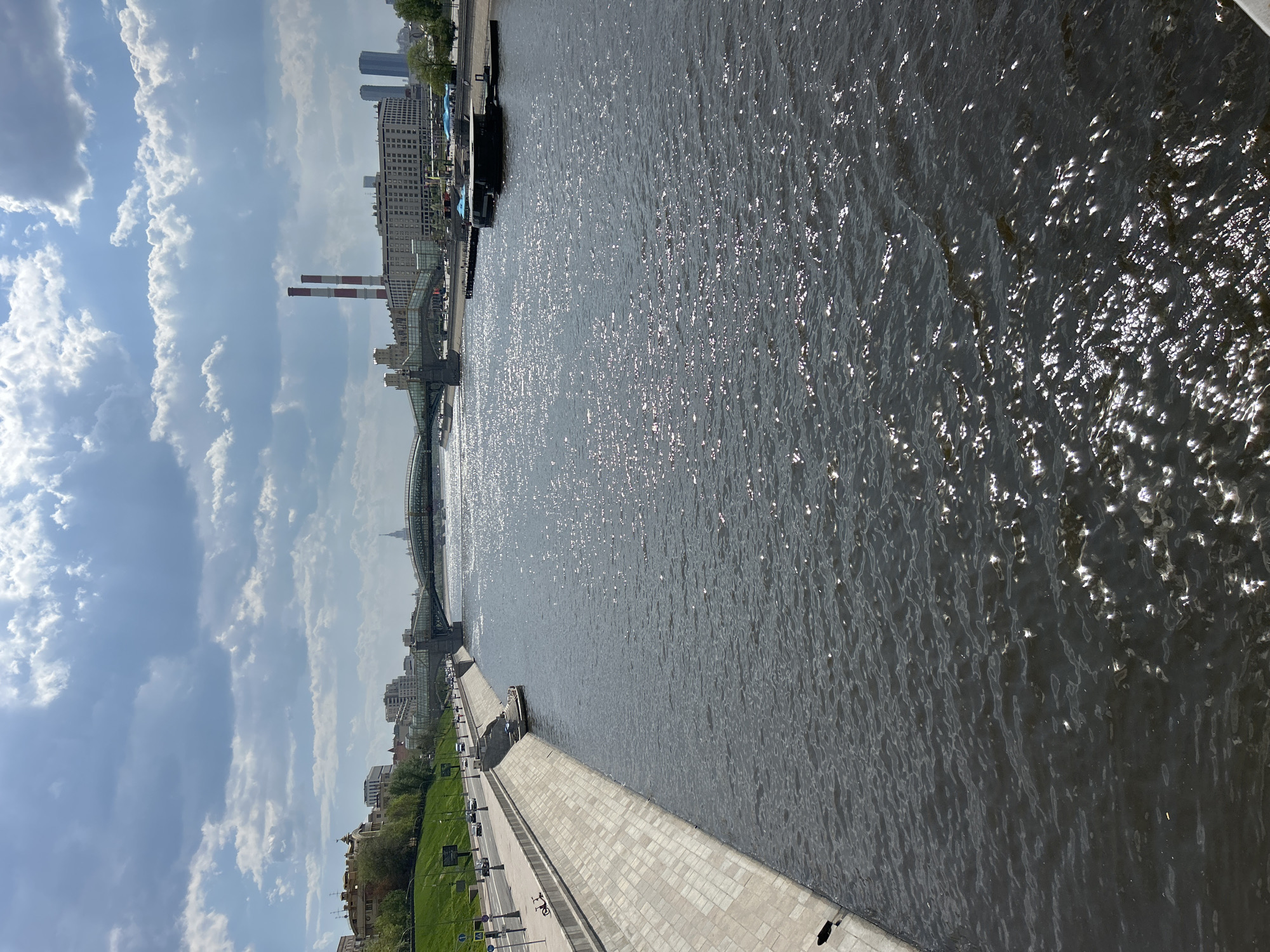 A view of a river in an urban setting. A bridge spans the river with buildings lining the riverbanks. Along the right side of the river, a stone embankment is visible, with a road running parallel. Buildings are situated along the riverbank in the background. Two tall chimneys are seen, and the sky is filled with cumulus clouds.