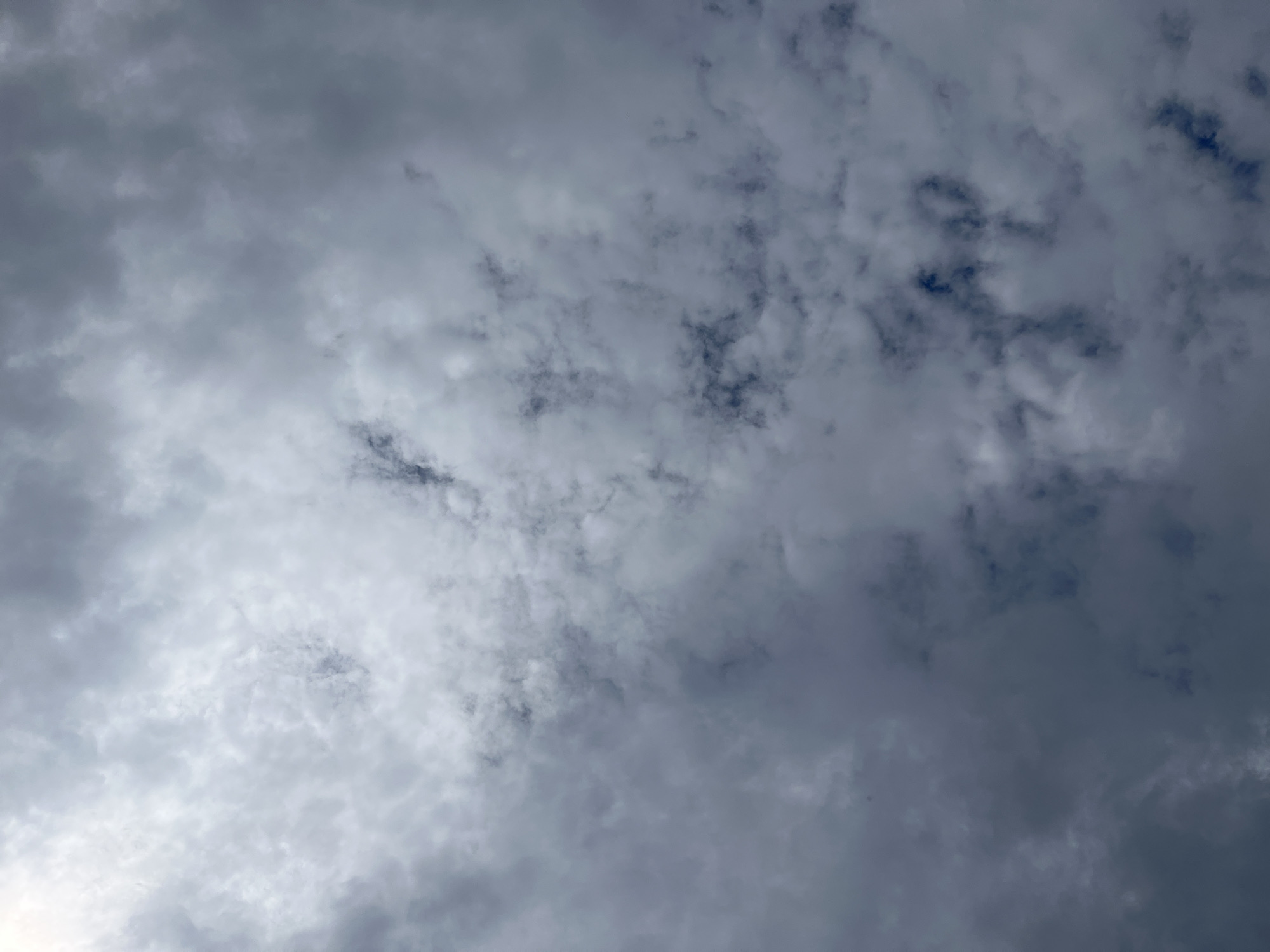 The image shows a sky filled with clouds. The clouds are varied in texture and density, ranging from light and wispy to dark and heavy. Patches of darker blue sky are visible through some of the thinner clouds, suggesting the presence of sunlight behind the overcast. The lighting indicates it is daytime, but the heavy cloud cover suggests overcast or potentially rainy weather. There are no discernible people, objects, or structures in the frame. The overall impression is a natural sky scene, likely taken from the ground looking upwards. No text is visible.