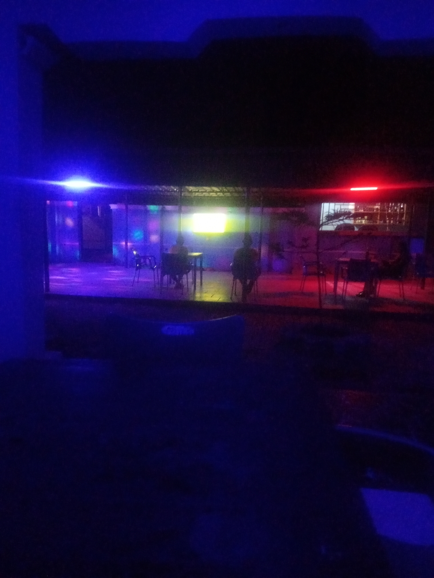 The image depicts an outdoor seating area of a business establishment at night, illuminated by a combination of blue and red lights, with disco-like colored lights inside. In the background, within an enclosed area, tables and chairs are visible. Two individuals are seated at separate tables; one is facing away from the camera, seated at a table with a chair. Another individual is also seated, facing away, near a table with chairs. The establishment appears to be a bar or restaurant in Sabon Gari, Nassarawa, Nigeria.