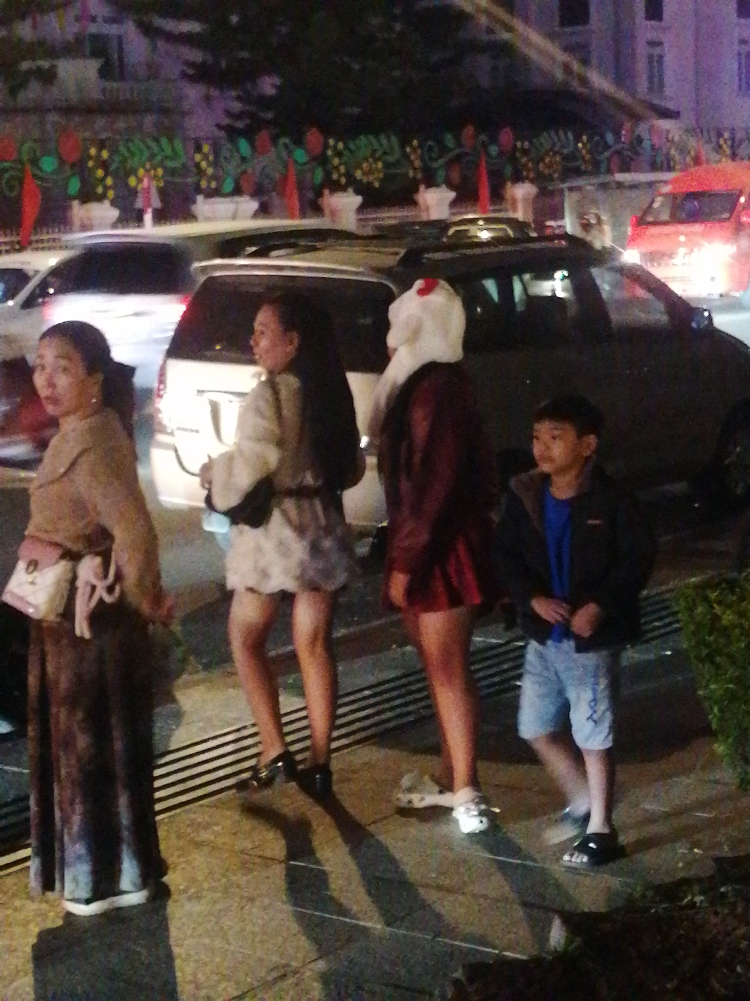 In Da Lat, Vietnam, four people are visible at night, seemingly outside. In the foreground to the left, a woman in a long, dark, patterned dress and a light brown top with a white and pink crossbody bag is facing left. Next to her, a woman in a short, white, textured dress and a dark belt is facing right. To the right of these two women, a young boy wearing a blue shirt and a dark jacket with grey trim is standing and looking to the left. Behind the boy, a person in a red dress and a white, full-body costume is partially visible. The background features several cars, including a red one, and decorative foliage along a wall. The ground is paved with concrete and appears wet, with faint horizontal lines suggesting a grate.