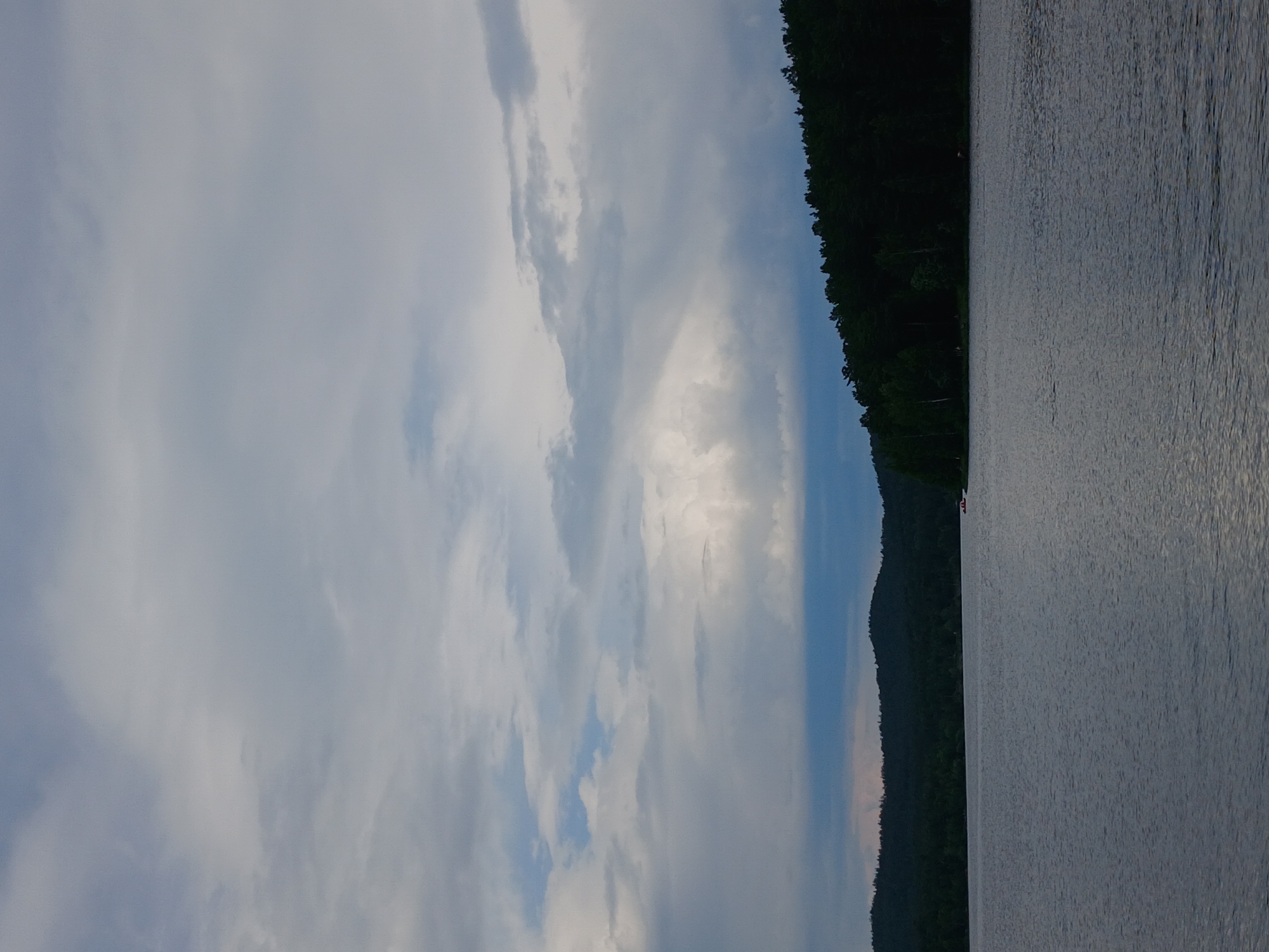 The image depicts a tranquil lake scene with a dark, forested shoreline under a cloudy sky. The water is calm, with subtle ripples reflecting the muted light. Hills covered in dense green trees form the backdrop, creating a sense of depth and wilderness. In the distance, a small red boat is visible on the water, adding a touch of human presence to the otherwise natural landscape. The sky is filled with a mix of gray and white clouds, with hints of blue peeking through, suggesting an overcast or potentially approaching weather system. The overall atmosphere is peaceful and serene, evoking a sense of natural beauty and quietude. The location is identified as the city of Revda, Russia, a place known for its natural landscapes and bodies of water, which align with the scene presented.