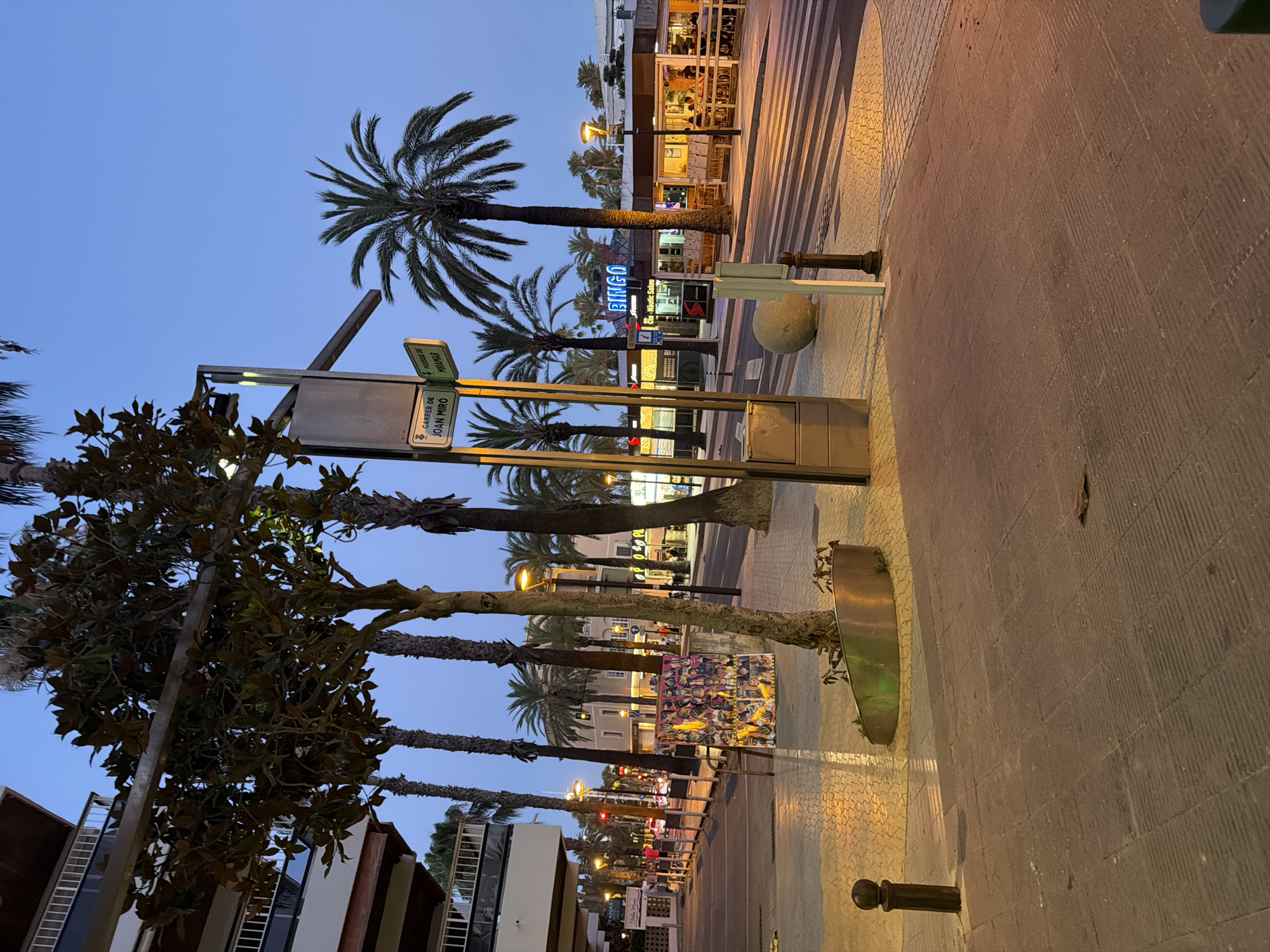 The image shows a street scene in Salou, Spain, likely during the early evening. A pedestrian walkway lined with palm trees and buildings runs along the right side of the image. In the foreground, the walkway is paved with textured grey tiles. A dark metal bollard is visible on the left, and a rounded metal planter contains a small tree. A street sign indicates "CARRER DE JOAN MIRÓ" and "PASSEIG DE MIRAMAR." Across the street, a crosswalk is illuminated by streetlights, casting long shadows. Several businesses with illuminated signs are situated along the street, including one with a "BINGO" sign. People are seated at outdoor tables in front of some establishments. A large, spherical object, possibly a stone decoration, is placed near the crosswalk. The sky is a clear, pale blue, indicating dusk.