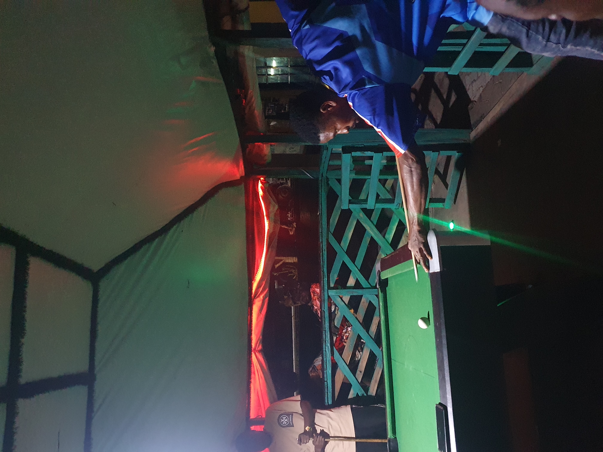 In Lokoja, Nigeria, at a dimly lit outdoor recreational area, two individuals are engaged in a game of pool. The foreground is dominated by a pool table with green felt and a single white ball positioned for play. A man, viewed from the side, is actively taking a shot, his arm extended and cue poised. He is wearing a blue and red athletic shirt and dark pants. To the left, another man, wearing a light-colored t-shirt and dark shorts, stands holding a pool cue, observing the game. The scene is set under a green tarp roof supported by dark beams, illuminated by red and green lights that cast a vibrant glow. Behind the pool table, a turquoise wooden barrier separates the playing area from the background, which is obscured by darkness but hints at further structures and possibly vehicles.