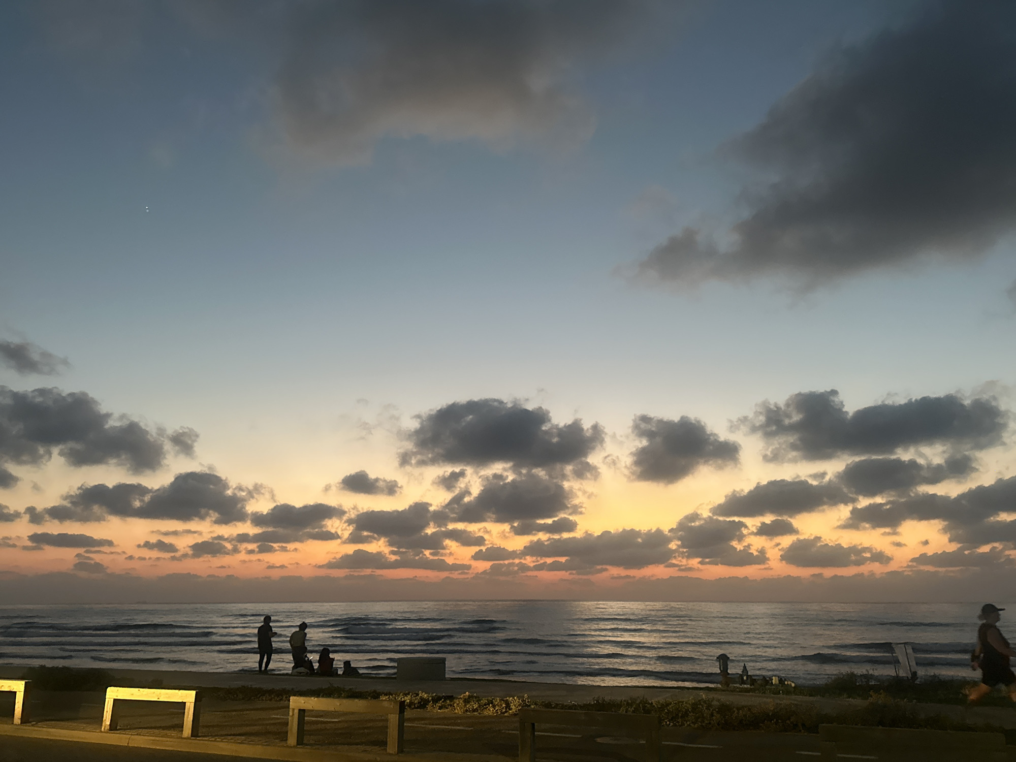 A wide-angle shot captures a serene evening seascape at a beach in Haifa, Israel. The sky transitions from a pale blue near the zenith to soft orange and yellow hues near the horizon, indicative of sunset. Wispy, dark clouds are scattered across the sky, with some denser formations present. The calm sea exhibits gentle waves rolling towards the shore. A silhouetted group of people stands on the beach, some appearing to be walking and others sitting. In the foreground, wooden barriers and some vegetation are visible, suggesting a promenade or public area. The overall lighting is soft and diffused, typical of twilight.
