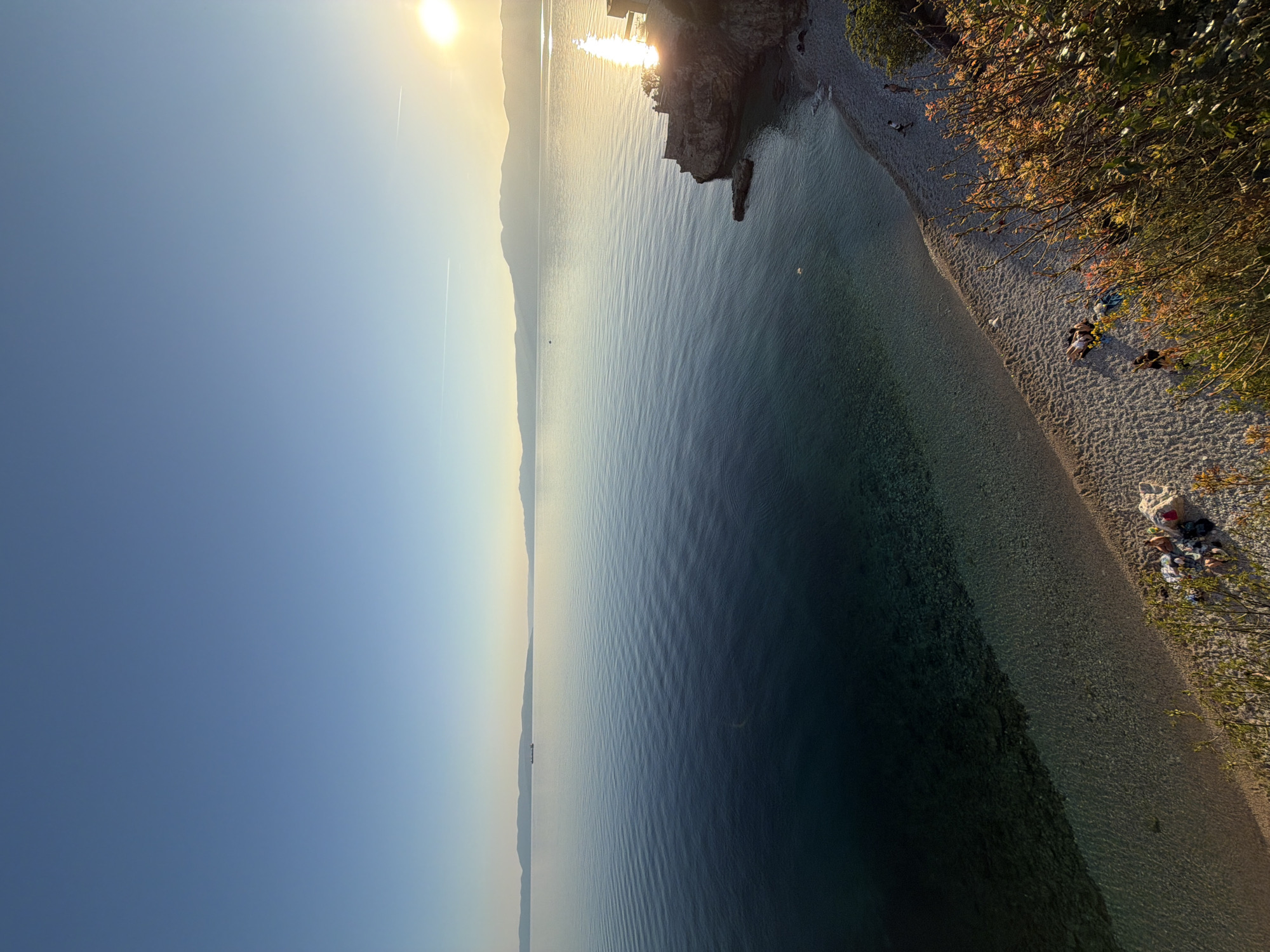 The image captures a scenic view of the Adriatic coast in Grad Rijeka, Croatia. The sun shines brightly, casting a glow across the water and sky. In the distance, the coastline stretches along the horizon. Closer to the viewer, a rocky outcrop juts into the sea. A narrow pebble beach lines the coast, and several people are relaxing or sunbathing near some trees. The water appears clear.