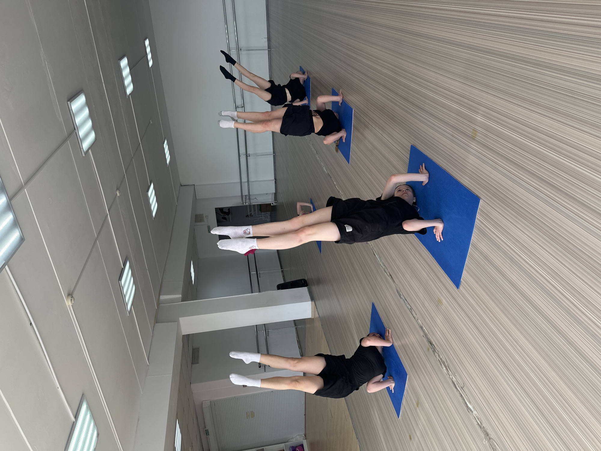 The image shows a group of four people in what appears to be a dance or exercise studio in Votkinsk, Russia. All individuals are lying face down on blue yoga mats, positioned in a line. They are dressed in black shirts and shorts, with varied choices of socks - some white, some black. Their legs are extended back and up. The studio has a wood-patterned floor and fluorescent lighting fixtures on the ceiling. A metal railing runs along the wall in the background.