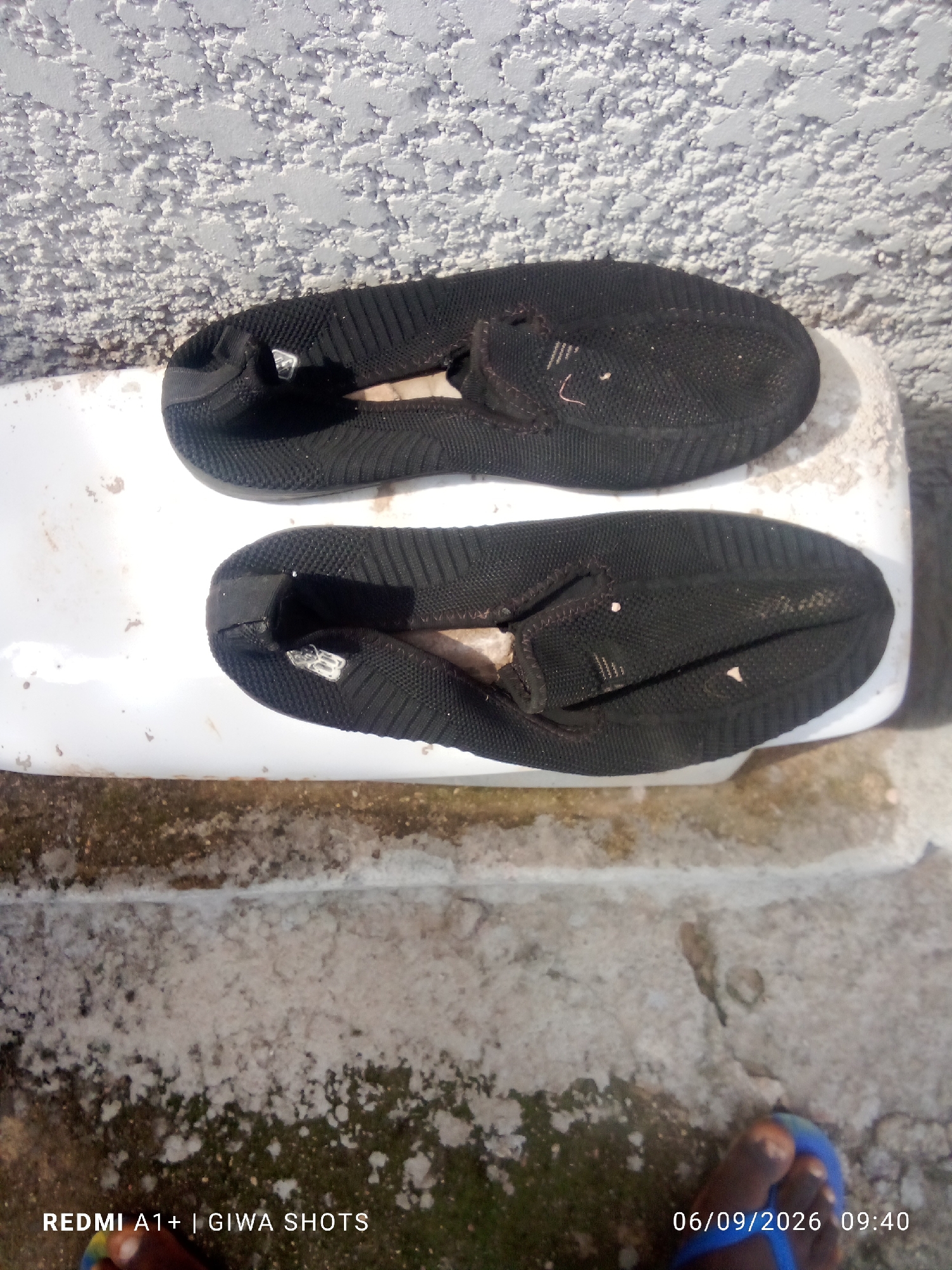 In Jalingo, Nigeria, a pair of black slip-on shoes rests on a white surface, possibly a ledge or furniture. The shoes are placed one on top of the other, with the top shoe slightly skewed. The texture of the white surface appears smooth, contrasting with the textured wall behind it, which has a rough, stucco-like finish in a light gray color. The ground below is a mix of concrete and dirt, showing patches of moss or dampness. The bottom of the image reveals a person's feet wearing blue flip-flops, partially visible. A timestamp and device information "06/09/2026 09:40 REDMI A1+ | GIWA SHOTS" are overlaid on the lower right and left corners of the image, respectively.