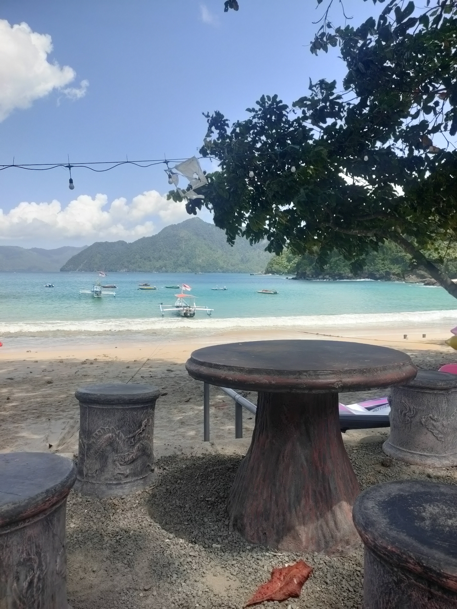 A scenic view of a tropical beach in Prigi, Indonesia, features a tranquil turquoise sea with gentle waves lapping onto a sandy shore. Several boats are anchored in the shallow waters, some adorned with red and white flags, while lush green hills rise majestically in the background under a clear blue sky dotted with fluffy white clouds. In the foreground, a sturdy table and matching cylindrical seats, adorned with intricate dragon carvings, are set on a bed of small grey pebbles, suggesting a casual seating area for beachgoers. A large tree with dark green foliage provides shade over the seating area, its branches extending towards the sky. The sand is scattered with small rocks and a single brown leaf.