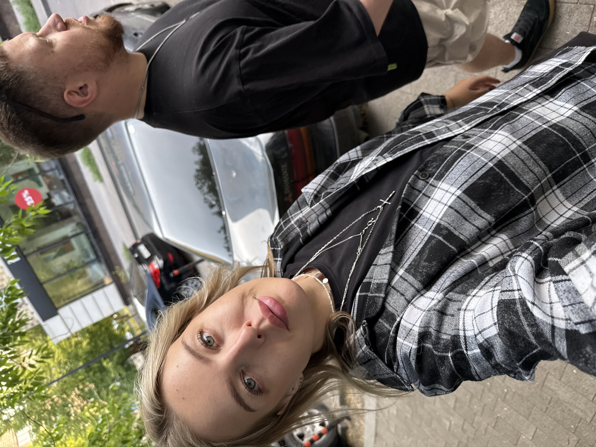A young woman with blonde hair and blue eyes, wearing a black and white plaid shirt over a black t-shirt and necklaces, smiles at the camera. She is standing on a sidewalk with a man with brown hair and a beard, wearing a black t-shirt and beige shorts, beside her. In the background, there are cars parked on a street, trees, and a building with storefronts. The scene appears to be a street in Krasnoyarsk, Russia, based on the context of the image.