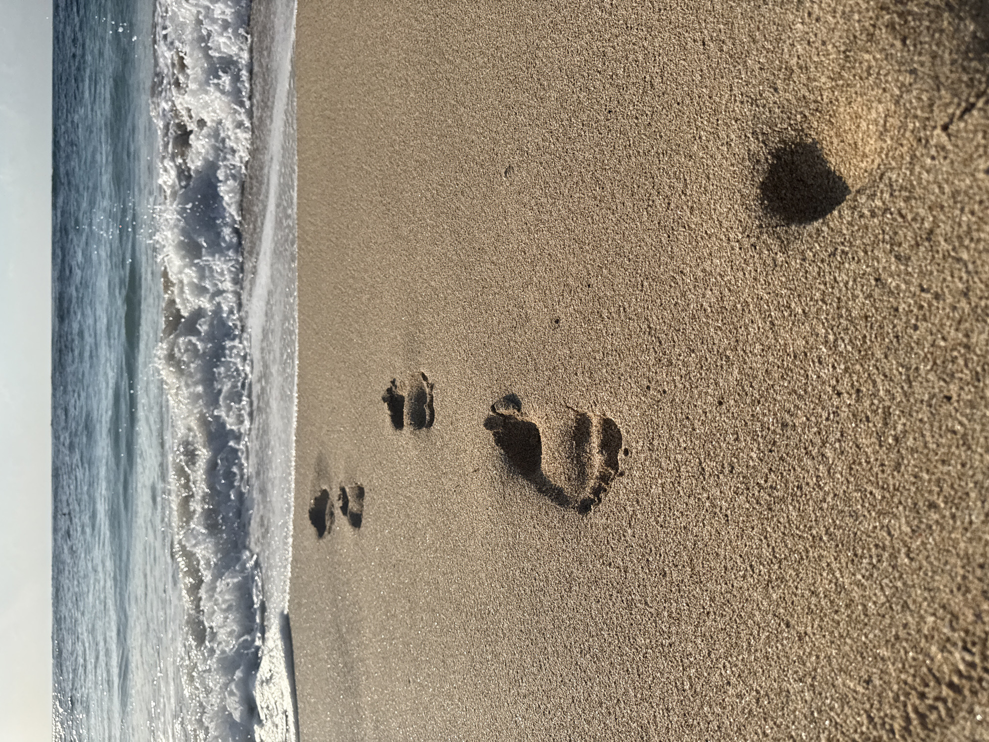 The image shows a close-up of footprints in the sand on a beach. The footprints are in a line leading away from the water. The ocean waves are breaking in the background, with blue water stretching to the horizon. The sand is light brown and textured, with some small pebbles visible. The lighting suggests it is daytime, with the sun casting shadows. The scene is likely Kuta Beach in Kuta Selatan, Indonesia, known for its sandy shores and ocean views.