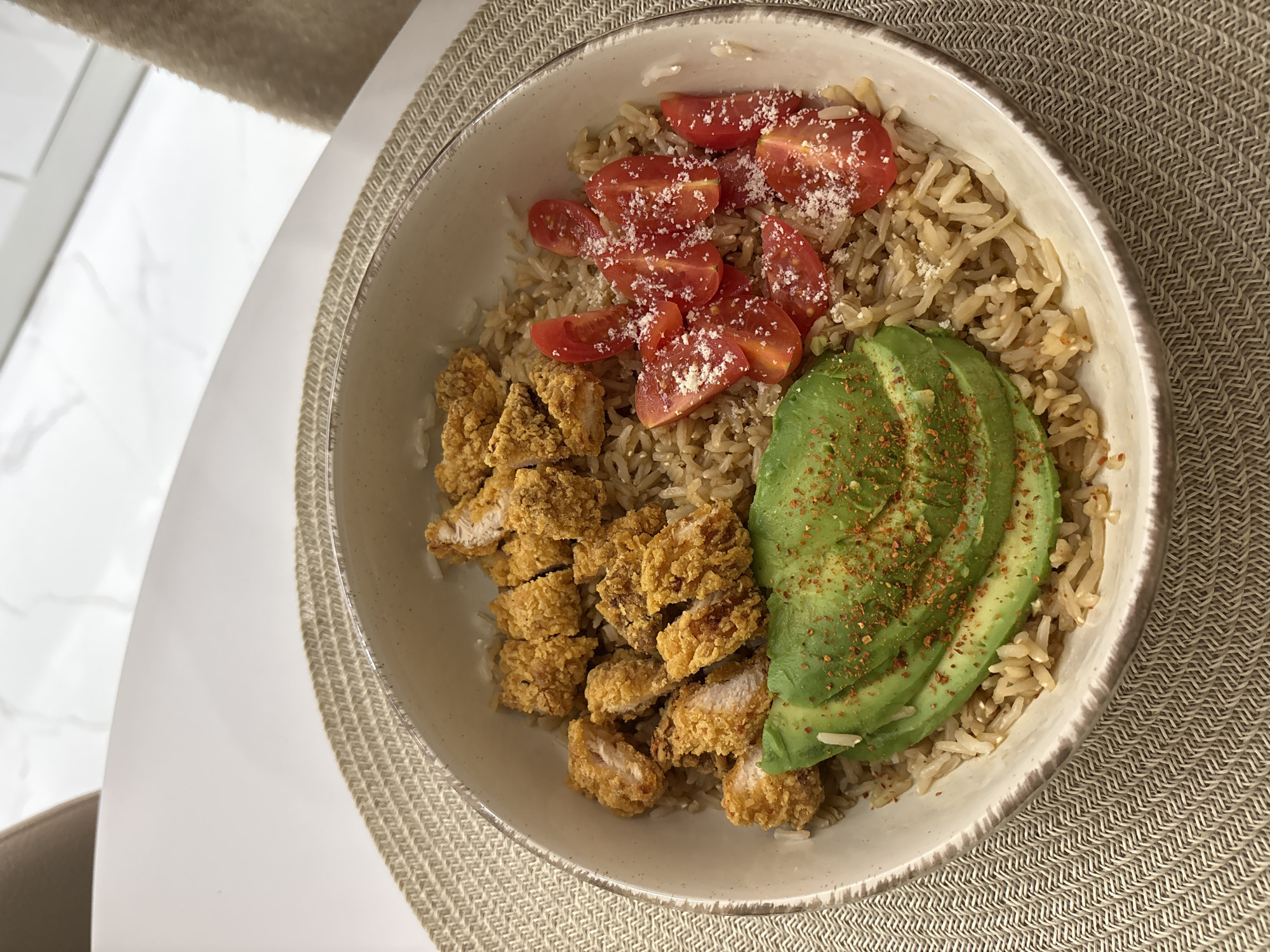 A high-angle, close-up shot depicts a meal within a ceramic bowl, resting on a woven placemat on a white surface. The bowl contains brown rice, sliced tomatoes topped with shredded cheese, pieces of breaded chicken, and avocado slices sprinkled with a reddish spice. The surface appears to be part of a table. The background shows a light-colored wall and a hint of a chair. There is no identifiable evidence linking this image to the city of Sofia, Bulgaria.