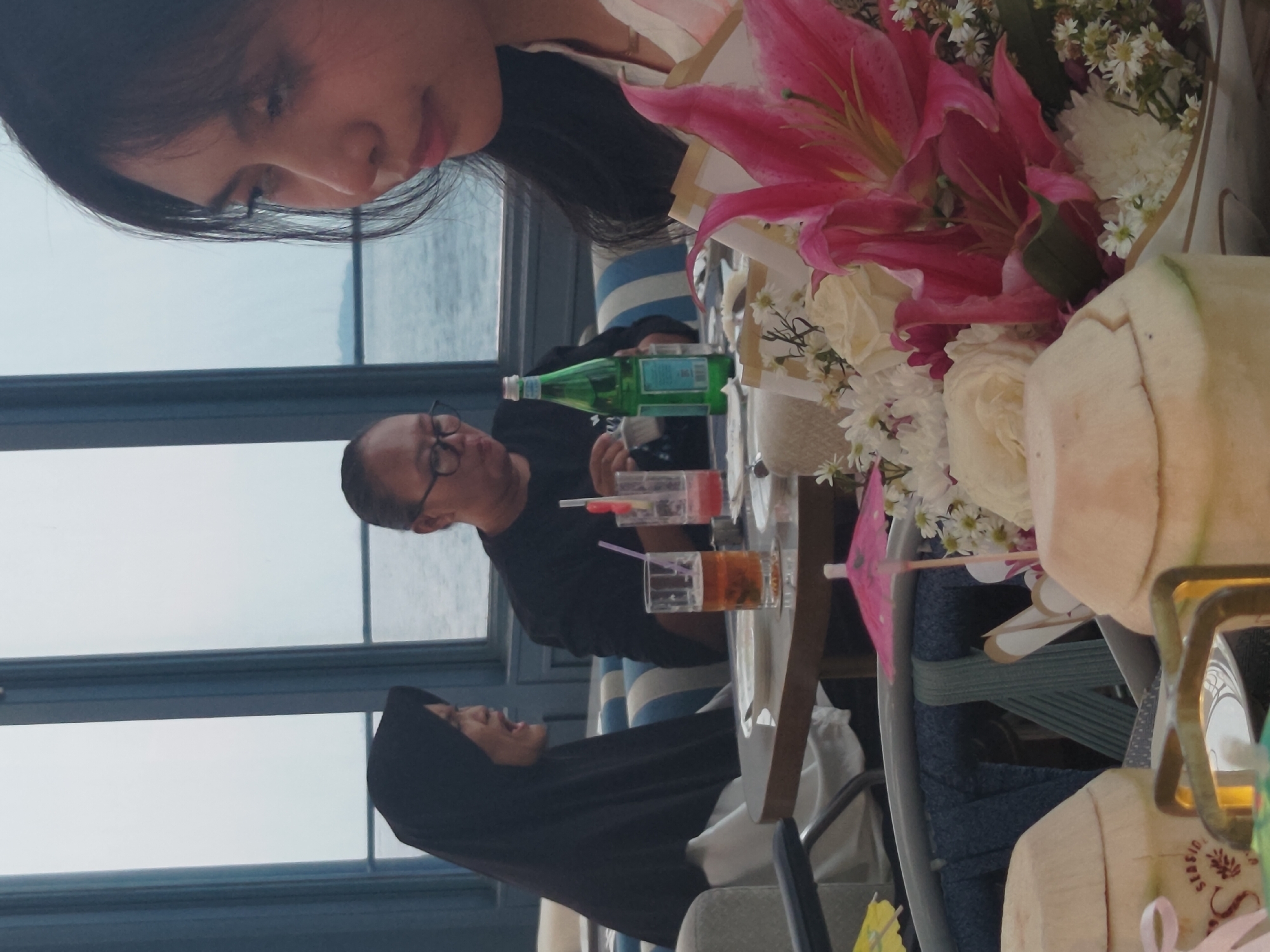 In a dimly lit restaurant with a view of the sea, three individuals are seated at a table. A woman wearing a black hijab is on the left, her mouth open as if speaking. In the center, another person with glasses, wearing a black shirt, is looking down. To the right, a young woman with dark hair is looking away from the camera. On the table, there are drinks, a green bottle, and a bouquet of pink lilies and white flowers. Two halved coconuts are placed in the foreground. The restaurant appears to be overlooking the ocean, with large windows in the background.