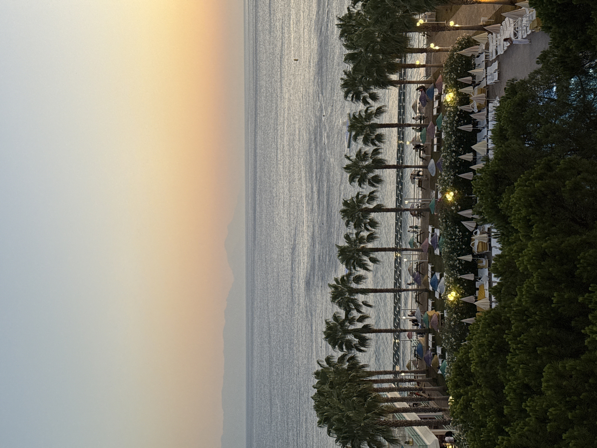 The image depicts a serene coastal resort scene at dusk, likely in Menderes, Turkey, given the style of architecture and vegetation. In the foreground, dense green pine trees create a lush border. Beyond them, a resort area is visible, featuring rows of empty lounge chairs and open umbrellas, suggesting a quiet evening. Several illuminated pathway lights are scattered throughout this area. Further back, a line of palm trees stands against the backdrop of a calm sea. In the distance, hazy mountains form a silhouette against the gradient sky, transitioning from a pale blue at the top to warm hues of orange and yellow near the horizon. The sea is a muted gray-blue, with subtle ripples indicating gentle waves. A few indistinct figures are present on the beach area behind the palm trees, some gathered near picnic tables under umbrellas. A yellow buoy is visible in the water. The overall atmosphere is peaceful and tranquil, with the soft lighting of dusk enhancing the natural beauty of the scene.