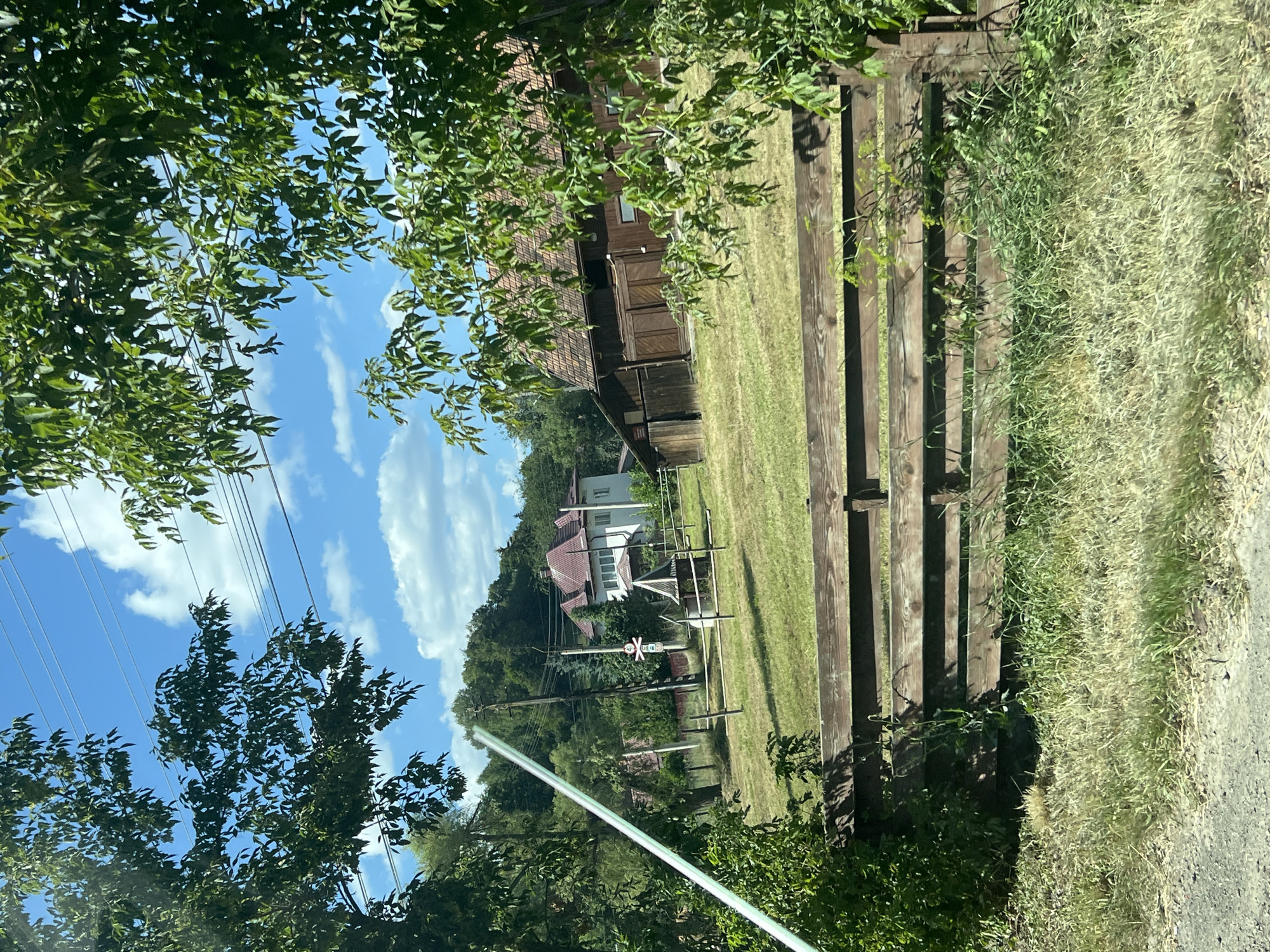 The image depicts a rural landscape in Molid, Romania, on a sunny day with a blue sky and scattered clouds. In the foreground, a weathered wooden fence runs horizontally across the frame, partially obscured by tall grass and foliage. Beyond the fence, a grassy field stretches out, leading to a traditional wooden barn with a tiled roof on the right side of the image. In the middle ground, a white house with a red roof is visible behind a cluster of trees, and further back, a densely wooded hill rises, dotted with utility poles and power lines. A white pole with a green stripe is leaning into the frame from the left, and a small, pyramid-shaped structure with a metal roof stands in the field near the house. The overall impression is one of a peaceful, rural setting.
